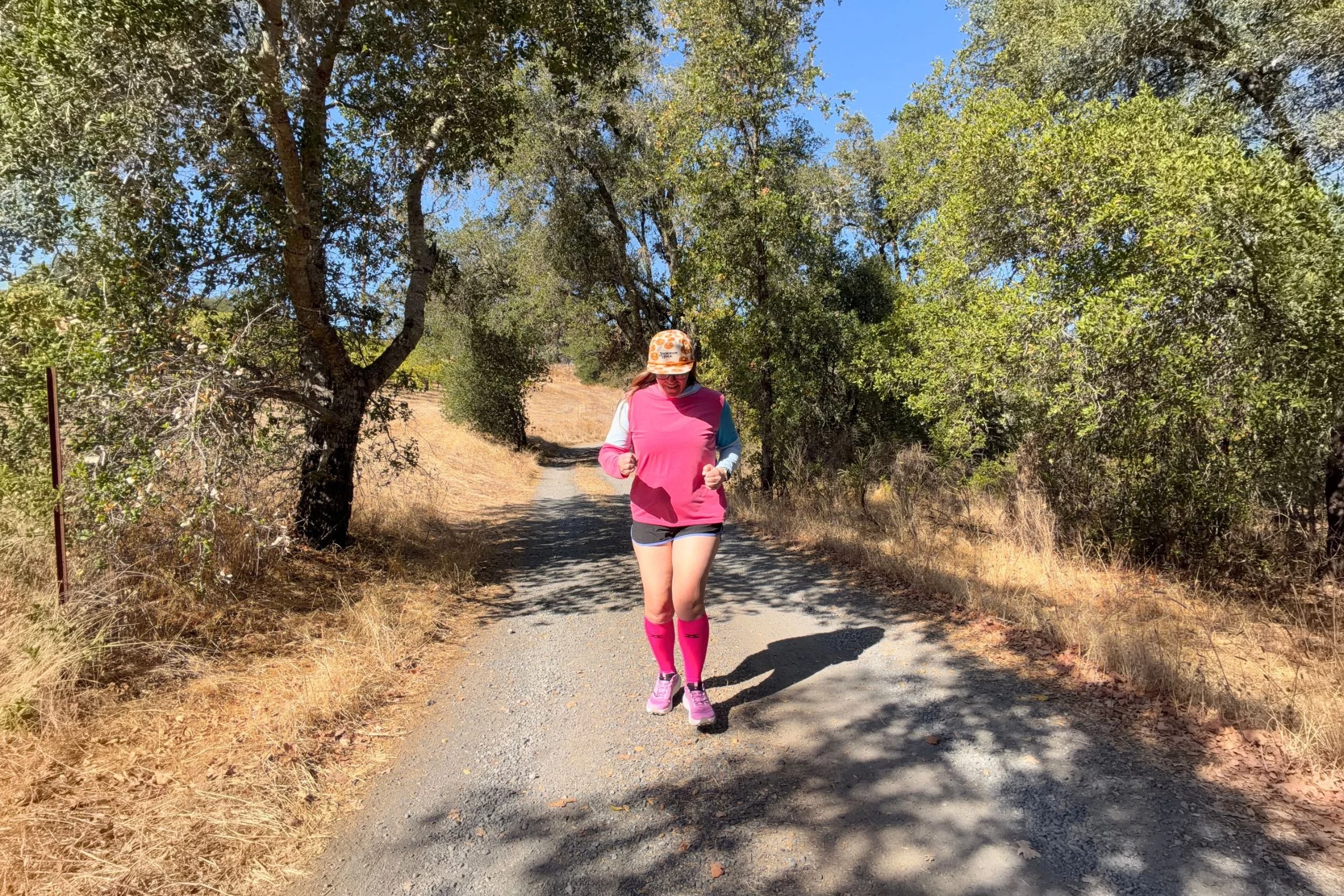   A runner wears bright pink Zensah Tech+ Graduated Compression Socks on a sunny, tree-lined trail, where their durable, tightly knit fabric helps protect against dirt and snags from brush.  