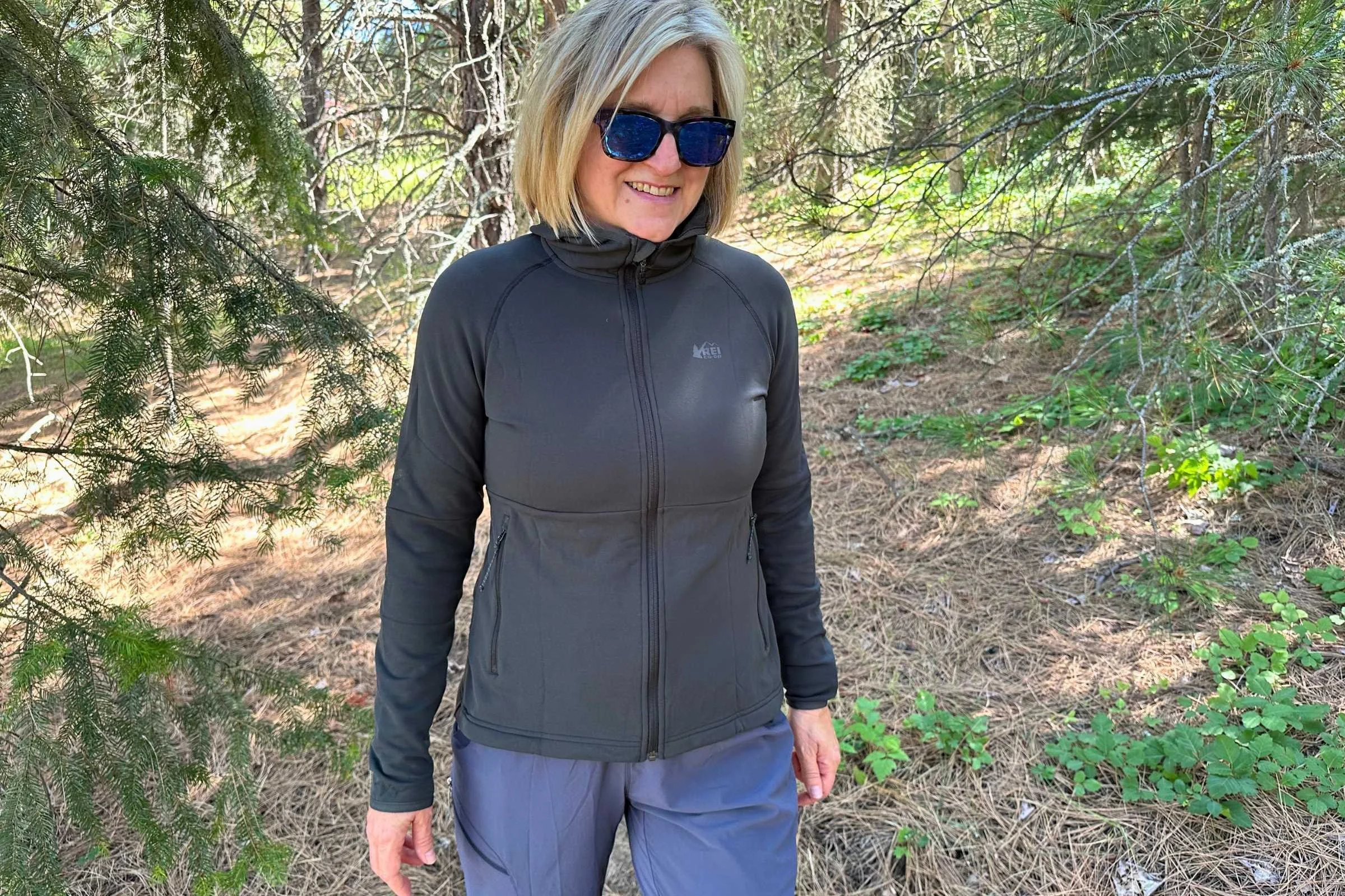 Naomi Hudetz wearing the black REI Co-op Flash Hyperstretch Fleece Jacket on a hike with trees in the background
