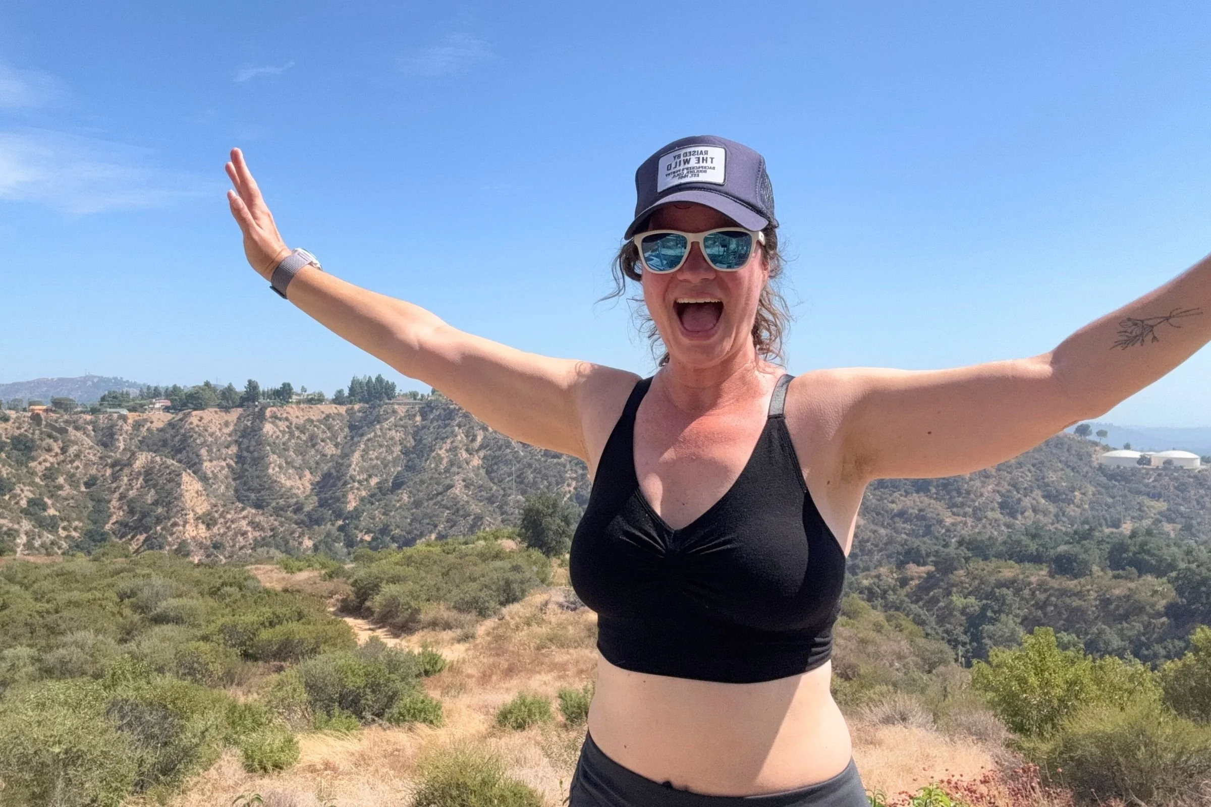 The author wearing the black BRANWYN Essential Supreme bra with her arms outstretched. Hills are in the background.