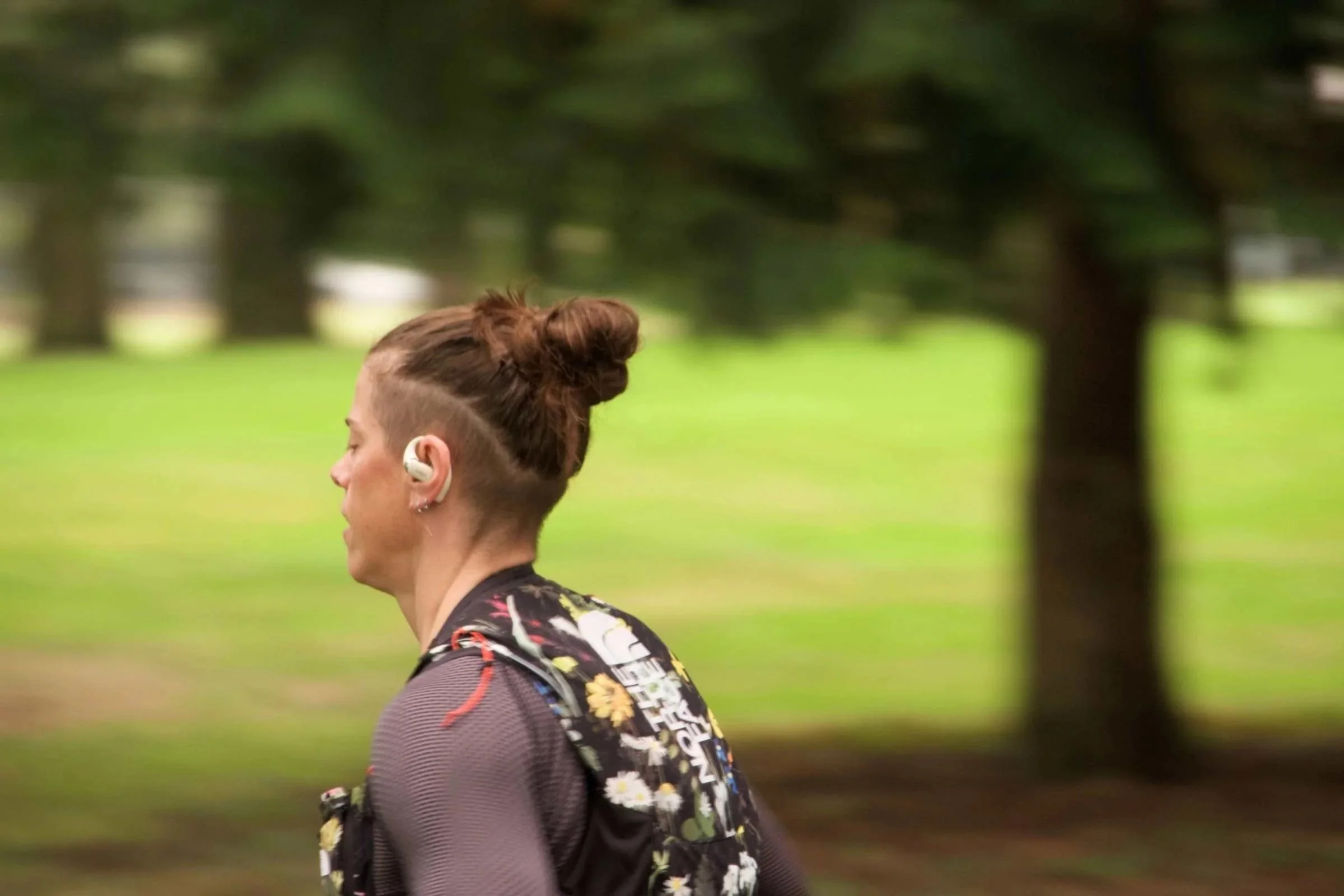 Person wearing the Shokz OpenFit 2 running headphones in a park with green grass