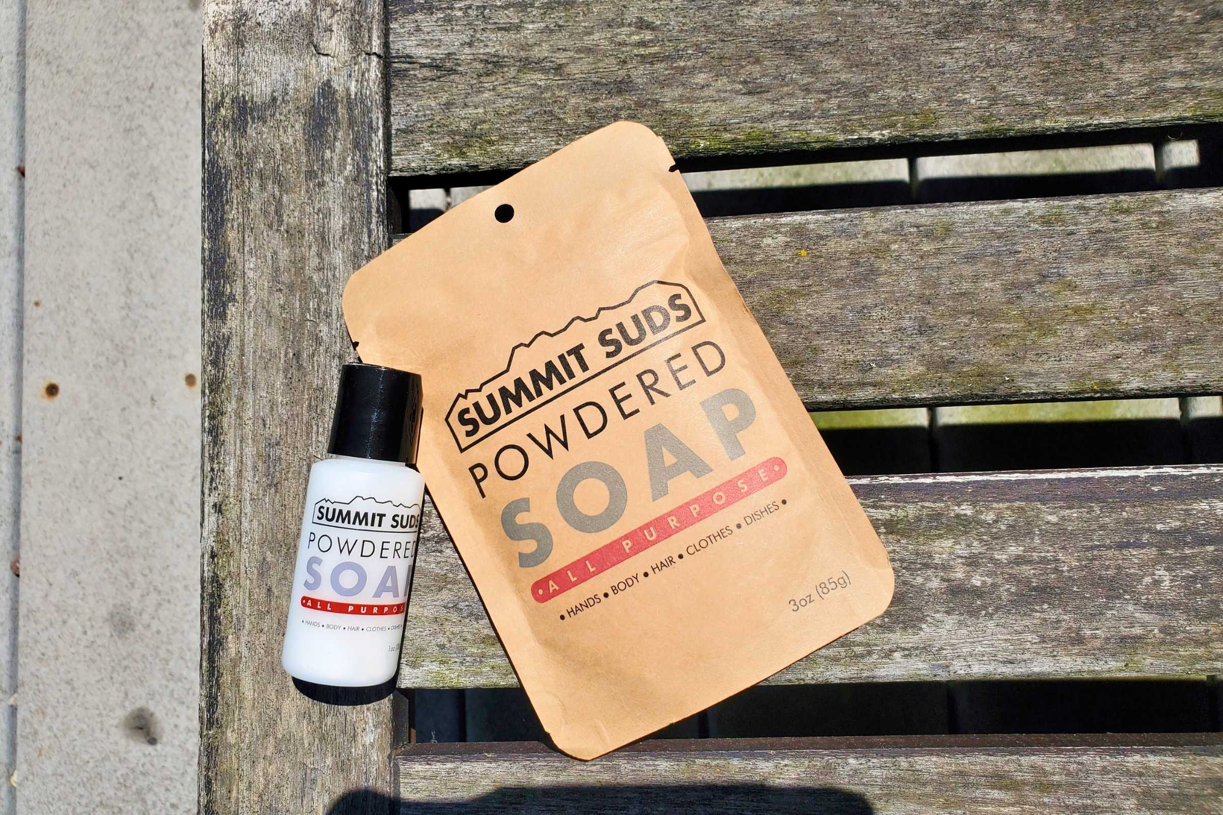 A small bottle of Summit Suds powdered soap next to a larger refill packet, lying flat on wooden lounge chair