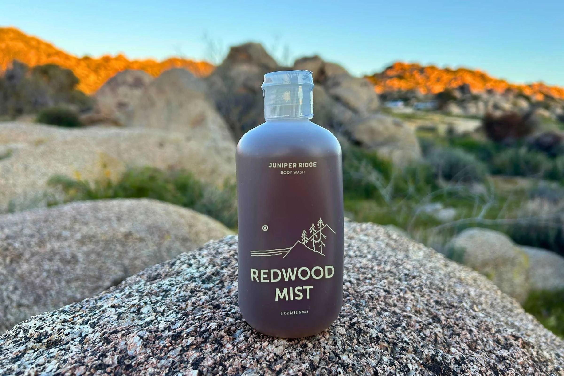 A brown bottle of Juniper Mist body wash in Redwood Mist scent sits on a speckled rock with rocks and hills in the background.