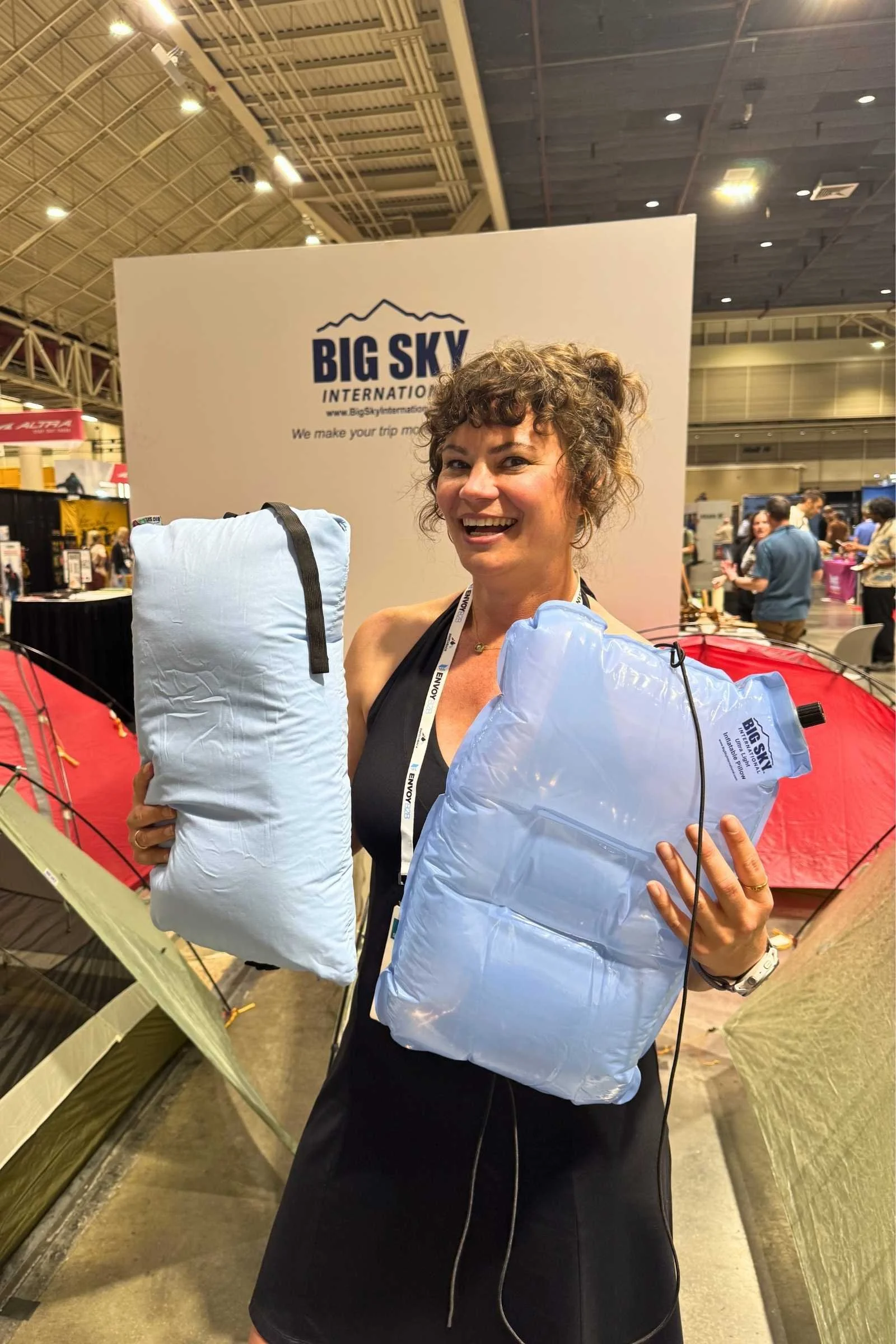A person holds two light blue camping pillows in front of a Big Sky International booth with tents in the foreground.