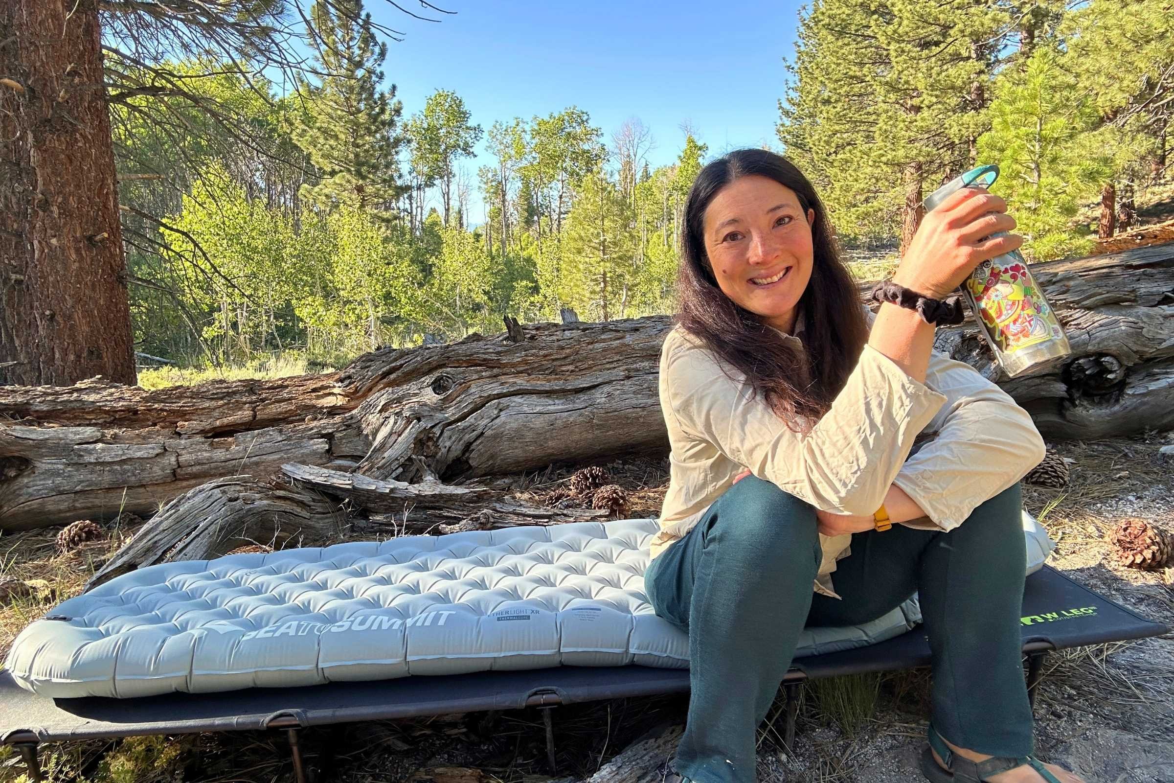 A person sits on a camping cot with Sea to Summit Ether Light XR Insulated Air Sleeping Pad in a forest