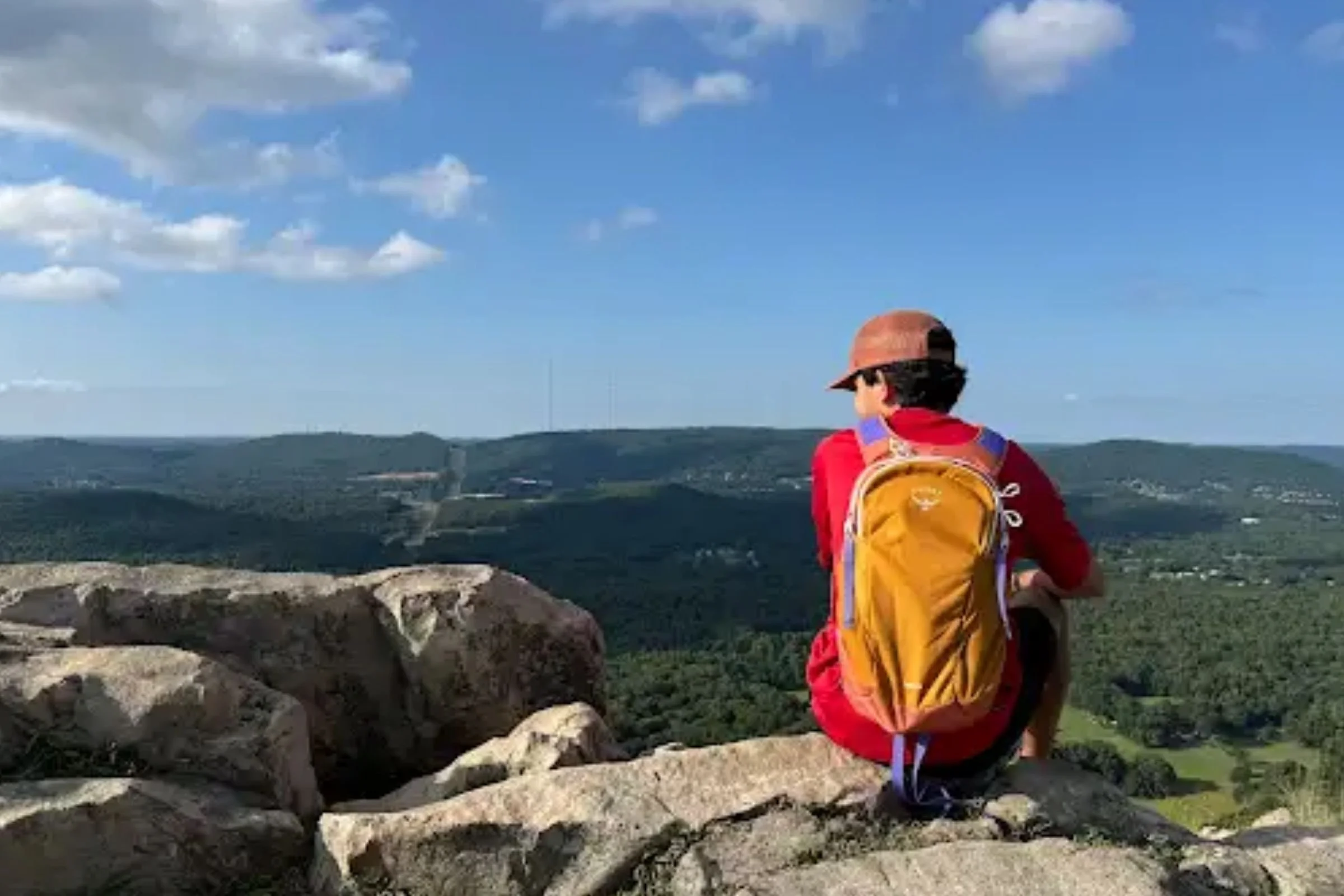 A person wearing the Osprey Daylite backpack while sitting atop a rocky outcropping with tree-covered hills in the distance.