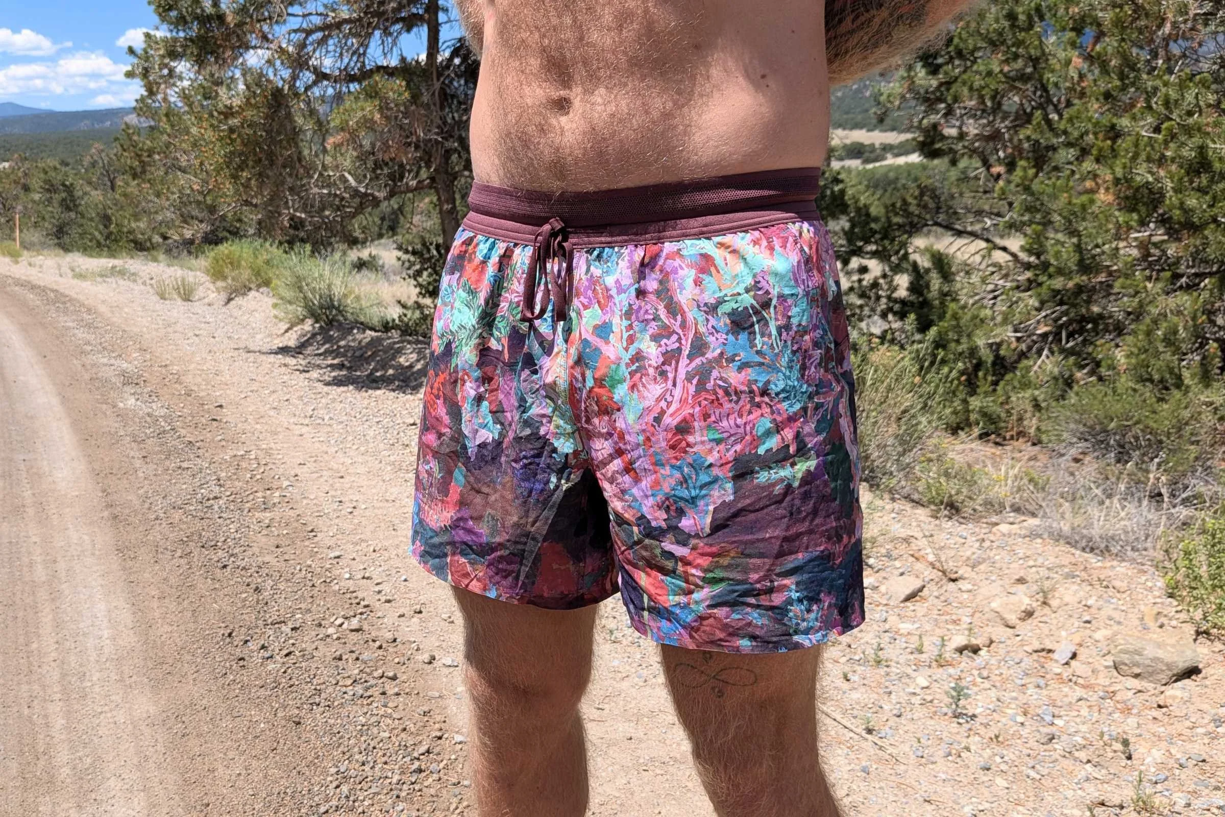 A man wearing Janji AFO Middle Running Shorts stands on a dirt road in a colorful outfit.