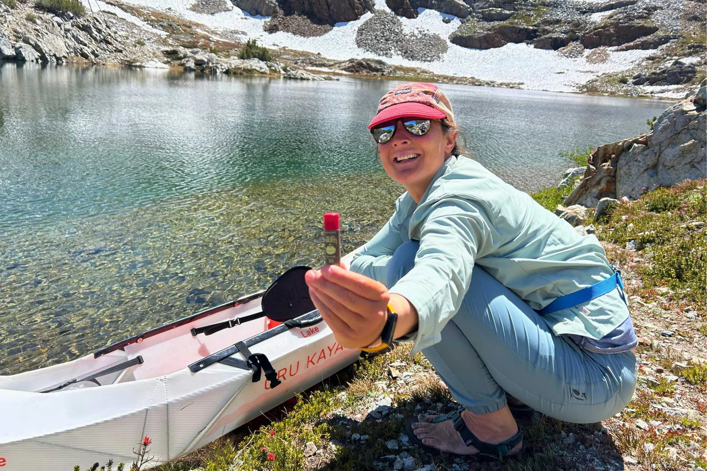 Tester holding Sun Bum SPF 30 Sunscreen Lip Balm beside alpine lake and kayak during high-elevation outdoor testing