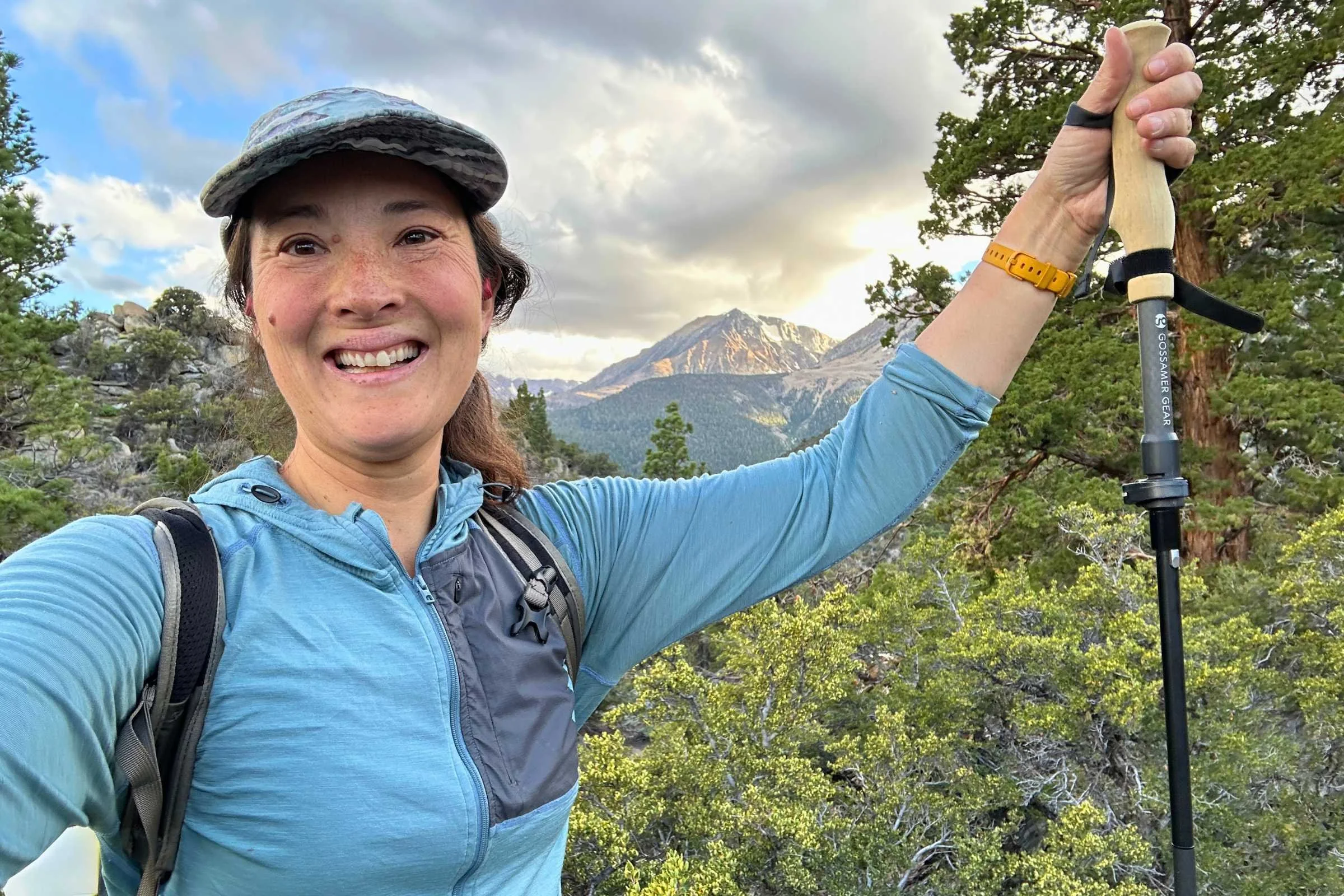 Author holding Gossamer Gear FT3 Folding Trekking Poles. In a wooded area with mountains in the background under a cloudy sky in the background.