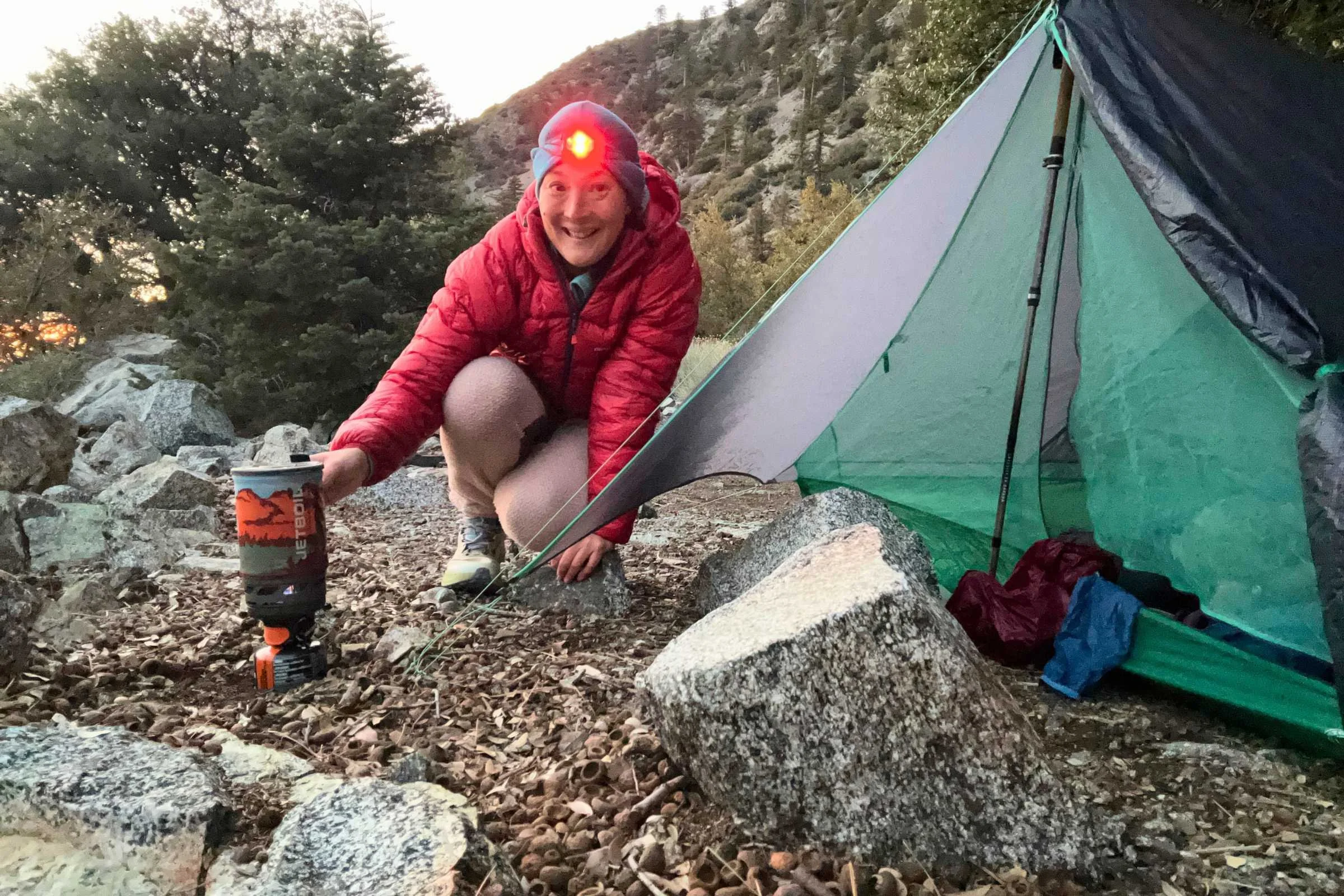 Backpacker wearing the Nitecore NU25 MCT headlamp in red-light mode while cooking beside a tent at sunrise.