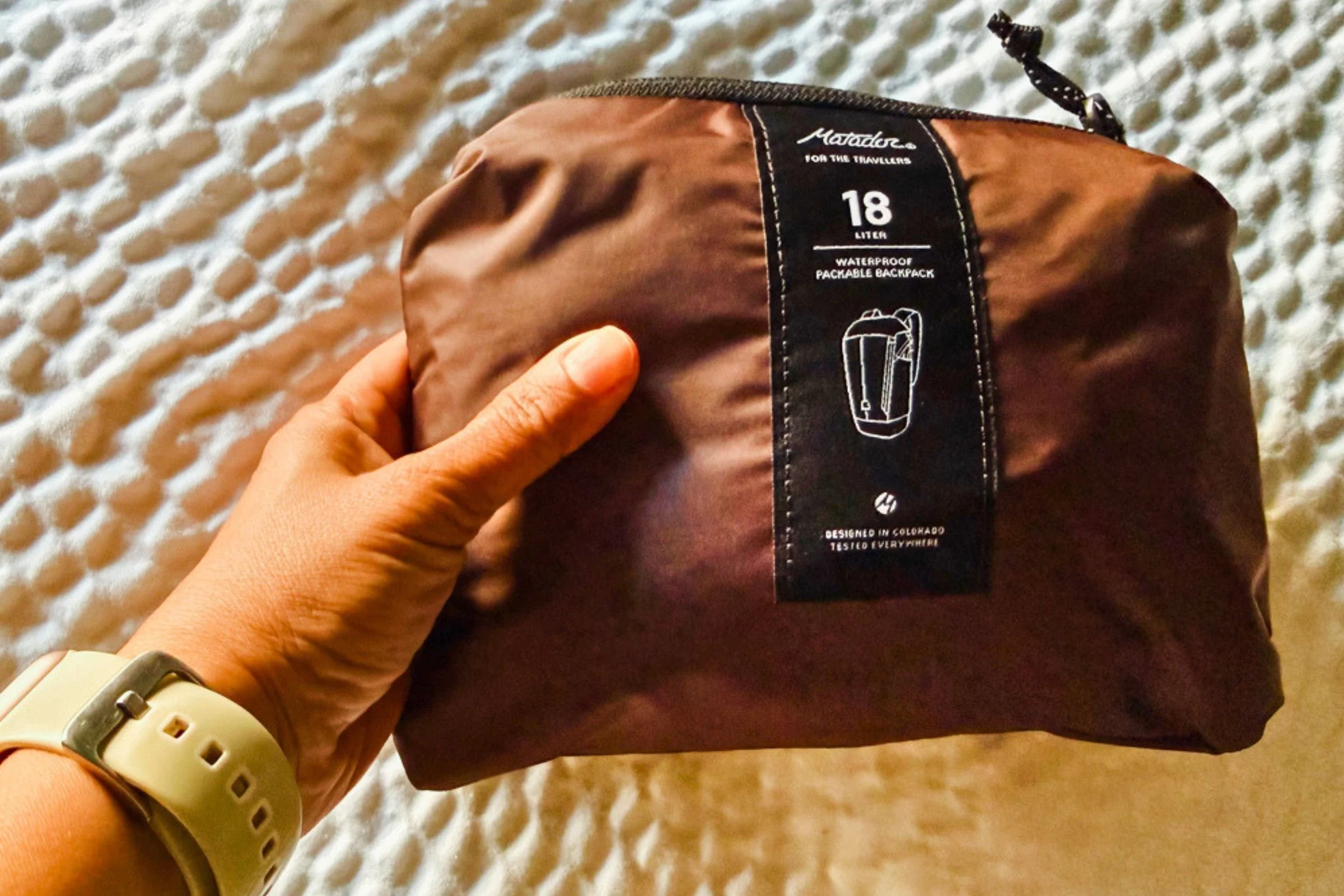 A hand holding the Matador 18L Waterproof Packable Backpack packed into its zippered pouch.