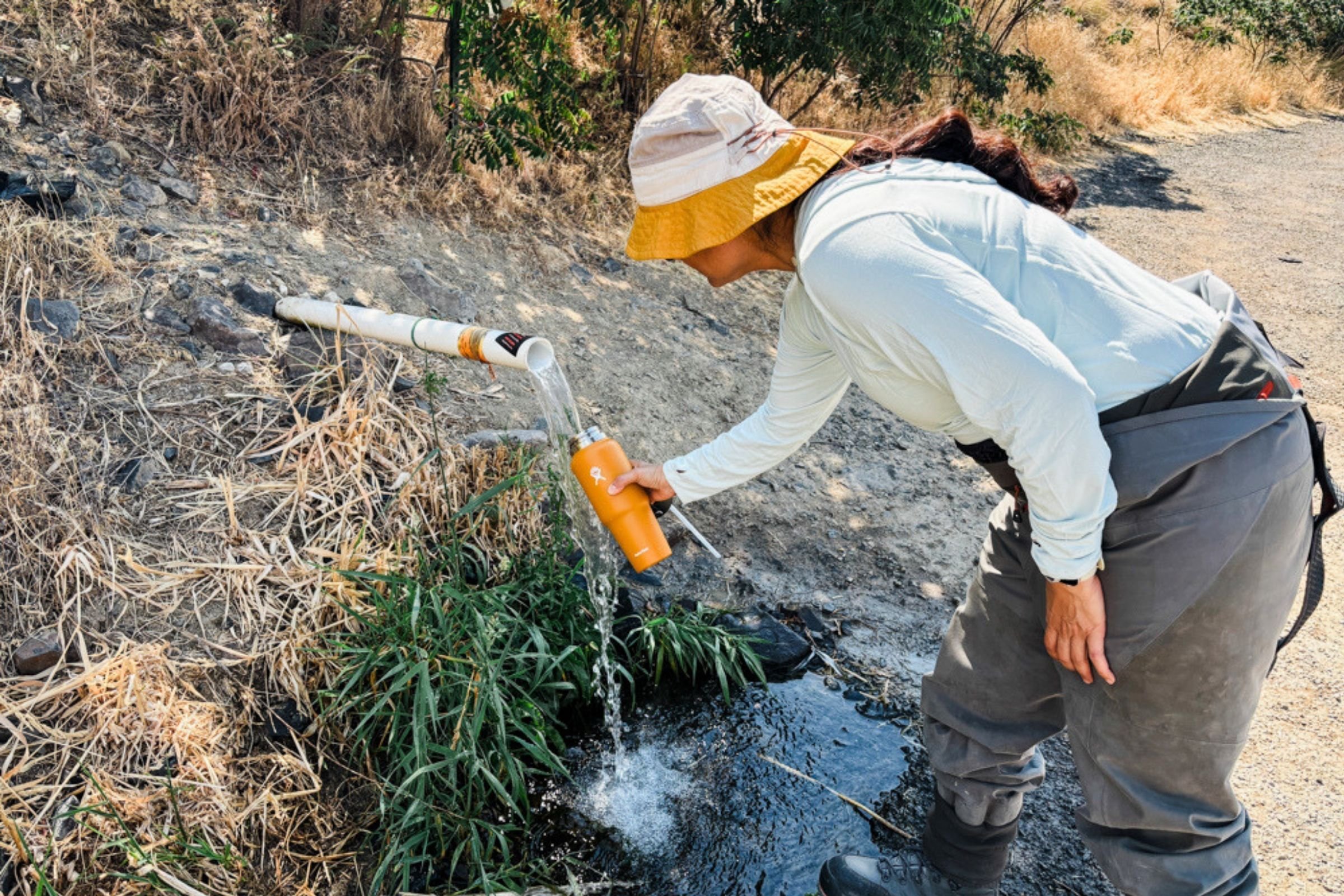 Woman in a hat and overalls filling a Hydro Flask 32 oz Travel Bottle from a stream.