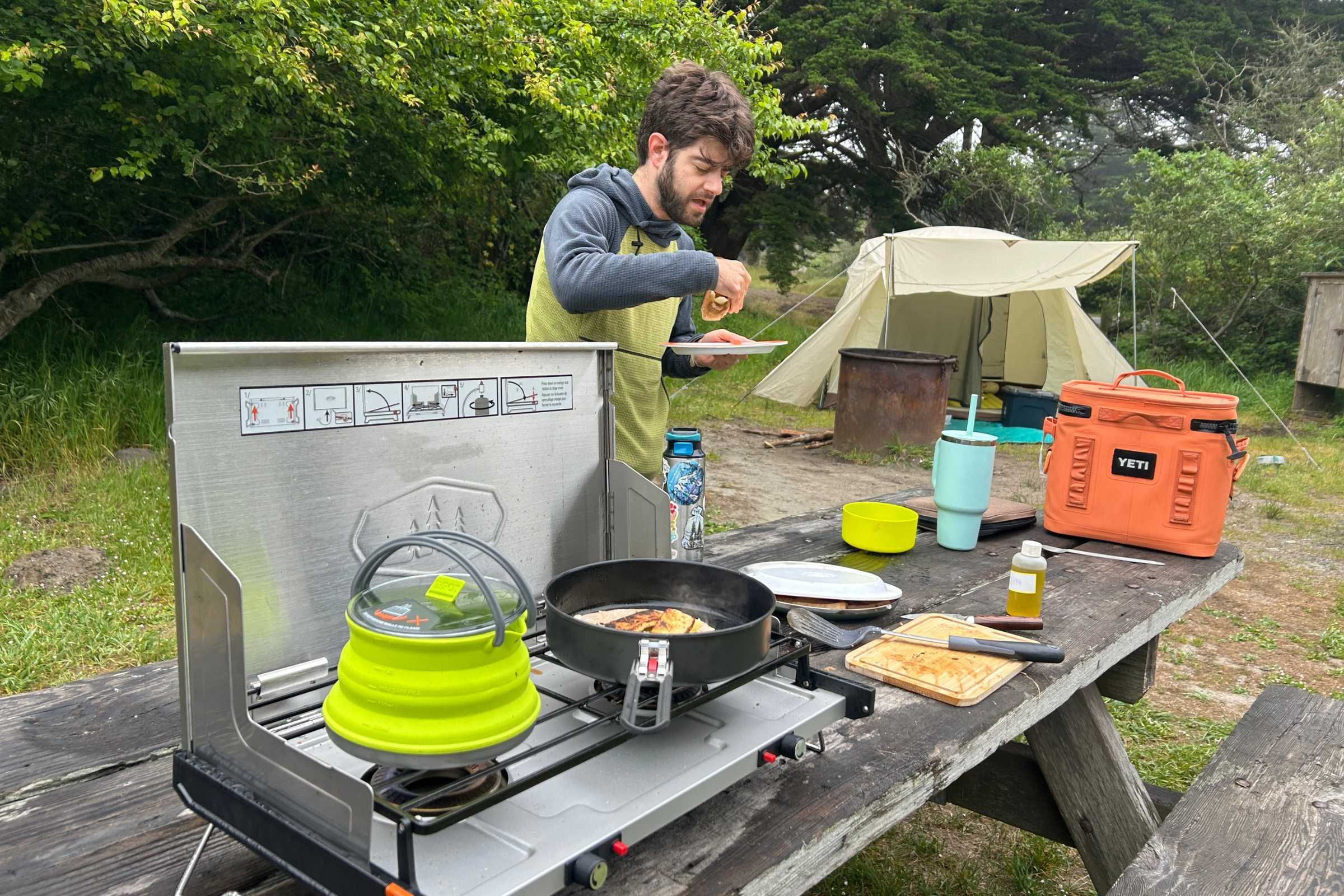 A person eating lunch standing next to the GSI Outdoors Pinnacle Pro 2-Burner stove on a wood picnic table.
