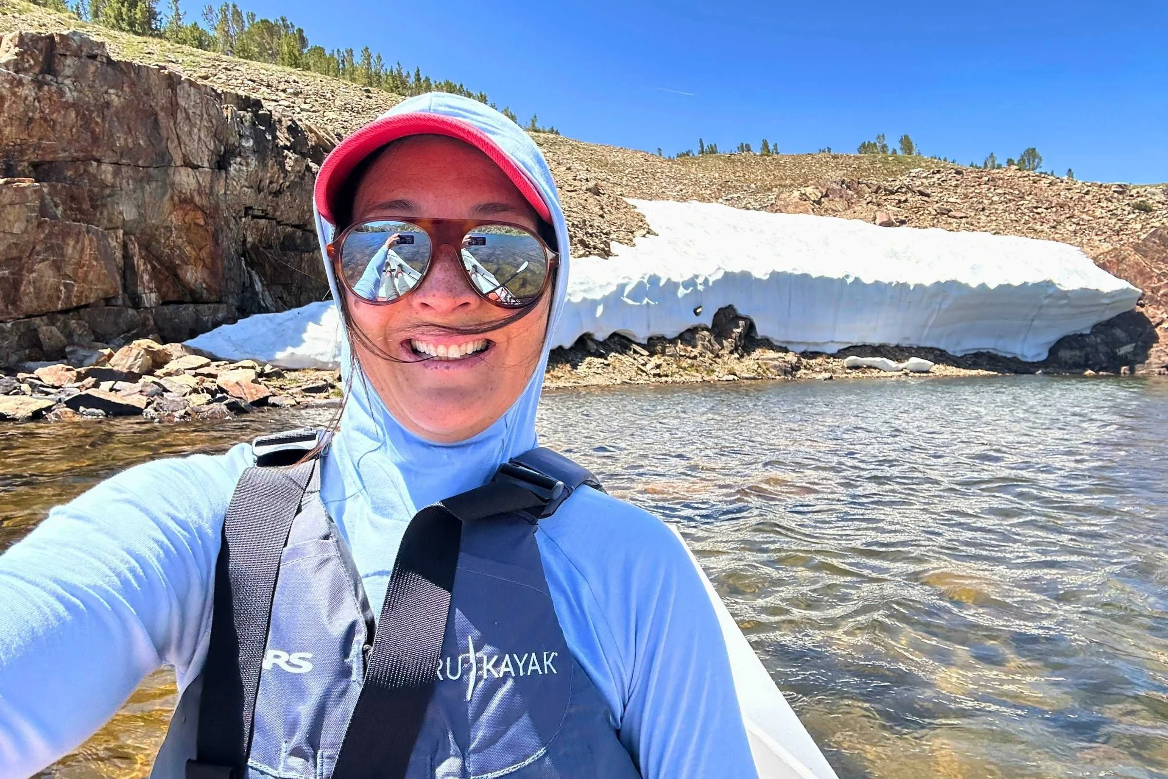 Woman kayak on an alpine lake with snow on the bank  while wearing a life jacket over a Free Fly Lightweight Hoodie with the hood up