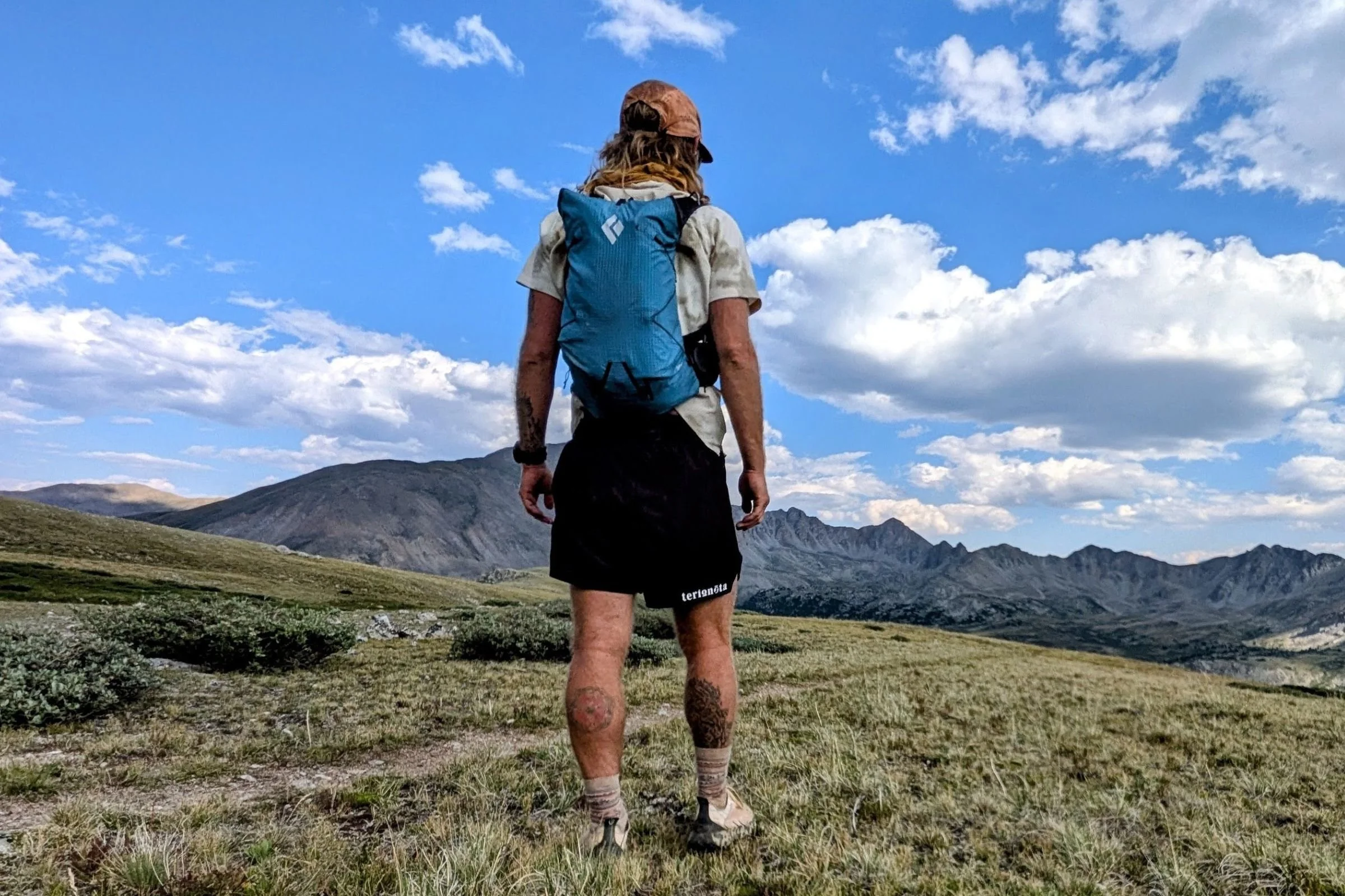 A man wearing the Black Diamond Distance 15 Backpack stands on a grassy hill.