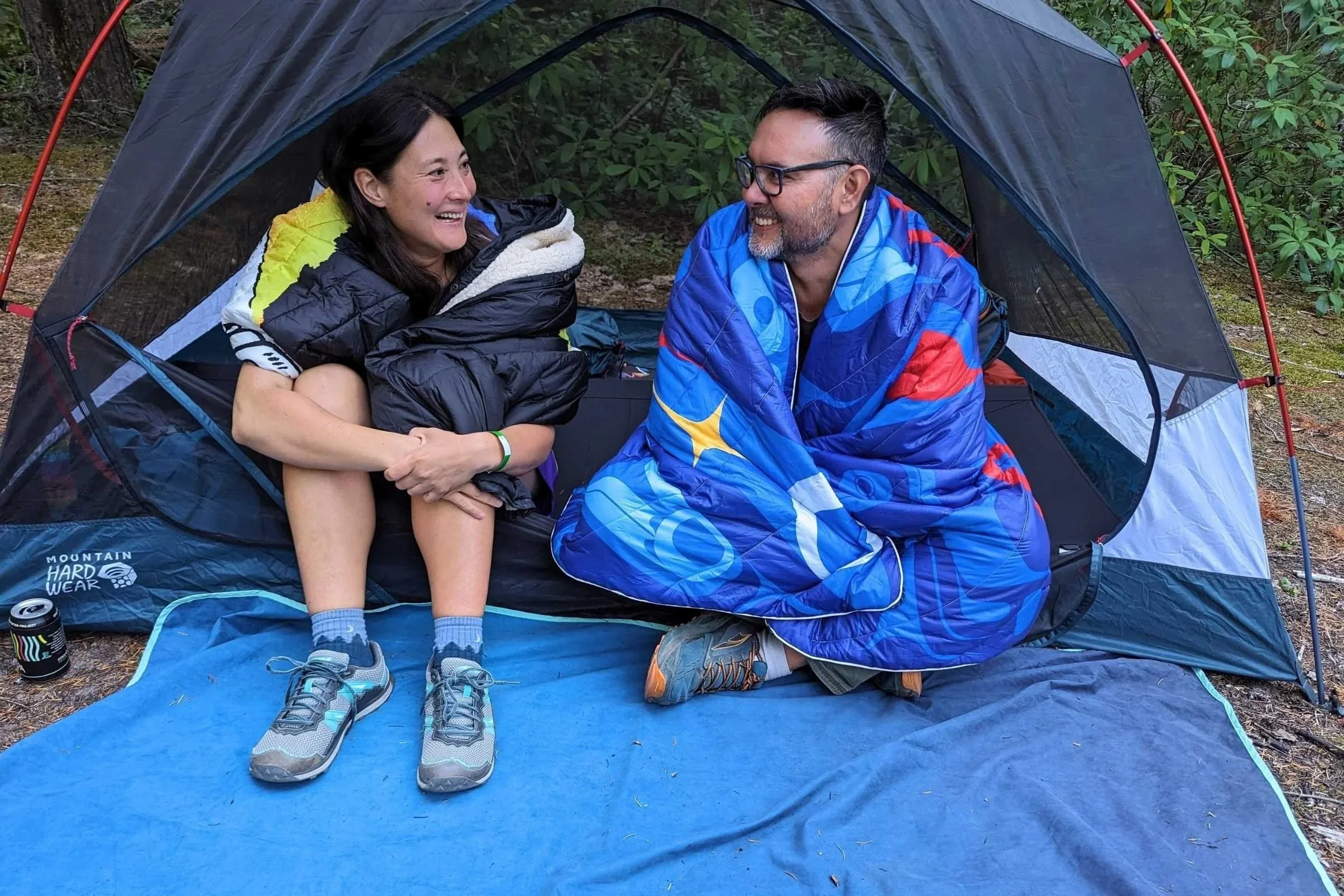 Two people sitting in a tent with camping blankets around their bodies