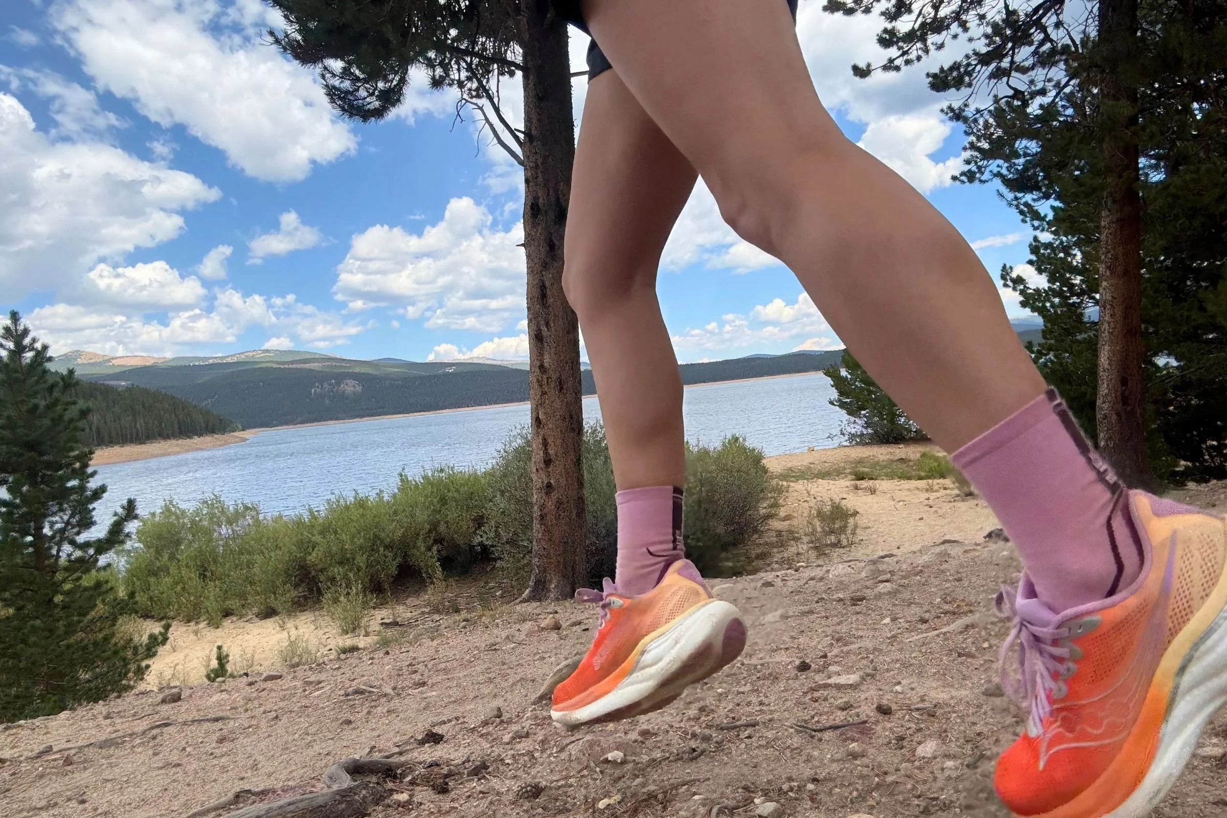 Orange Saucony Ride 19 Road Running Shoes and matching socks worn by a runner on a trail with a lake in the background.