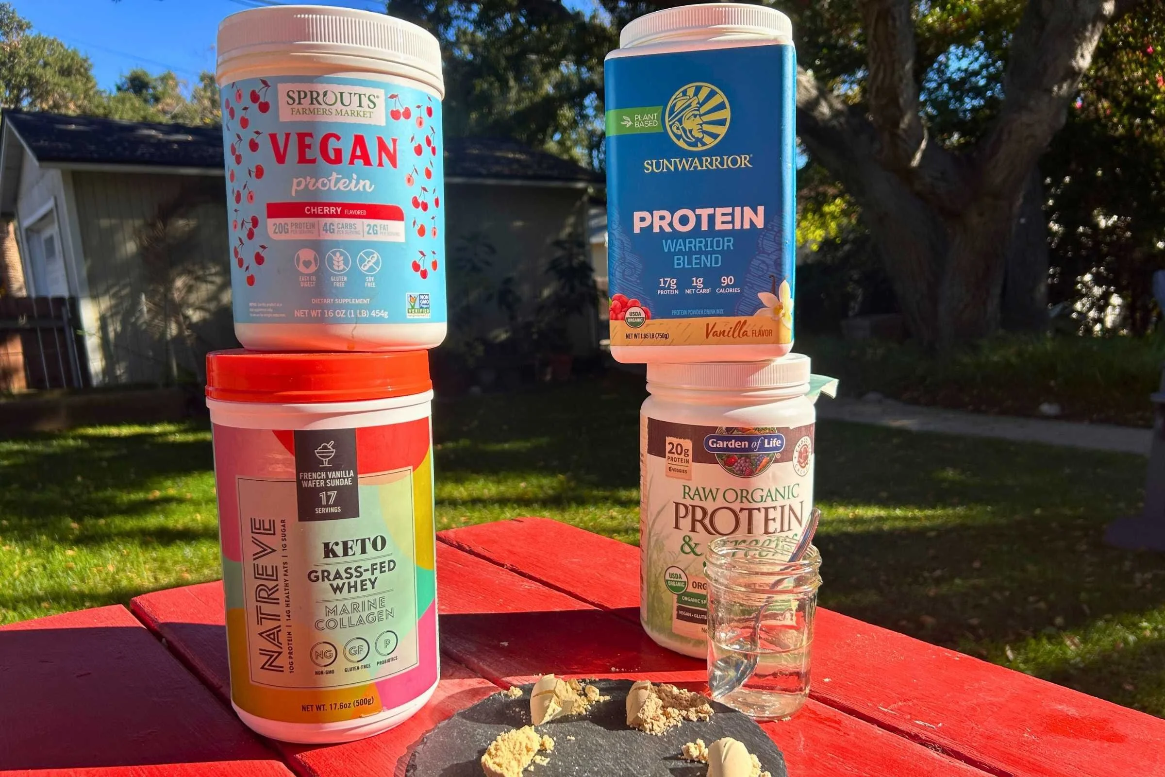 Four protein powder containers on a red picnic table with the powders displayed on a disc in front of them