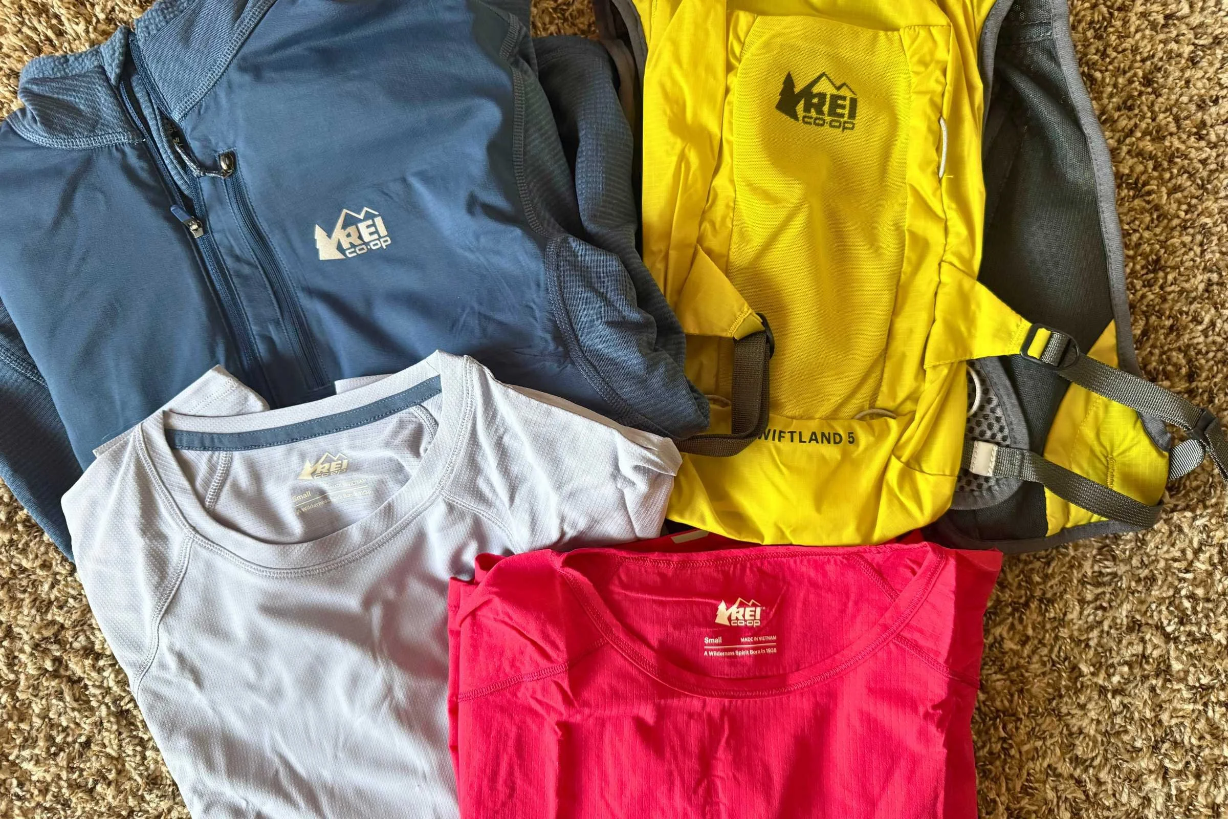 REI Co-op Swiftland running kit including Grid Running Shirt, jacket, and Swiftland 5 Hydration Vest laid out on the ground.