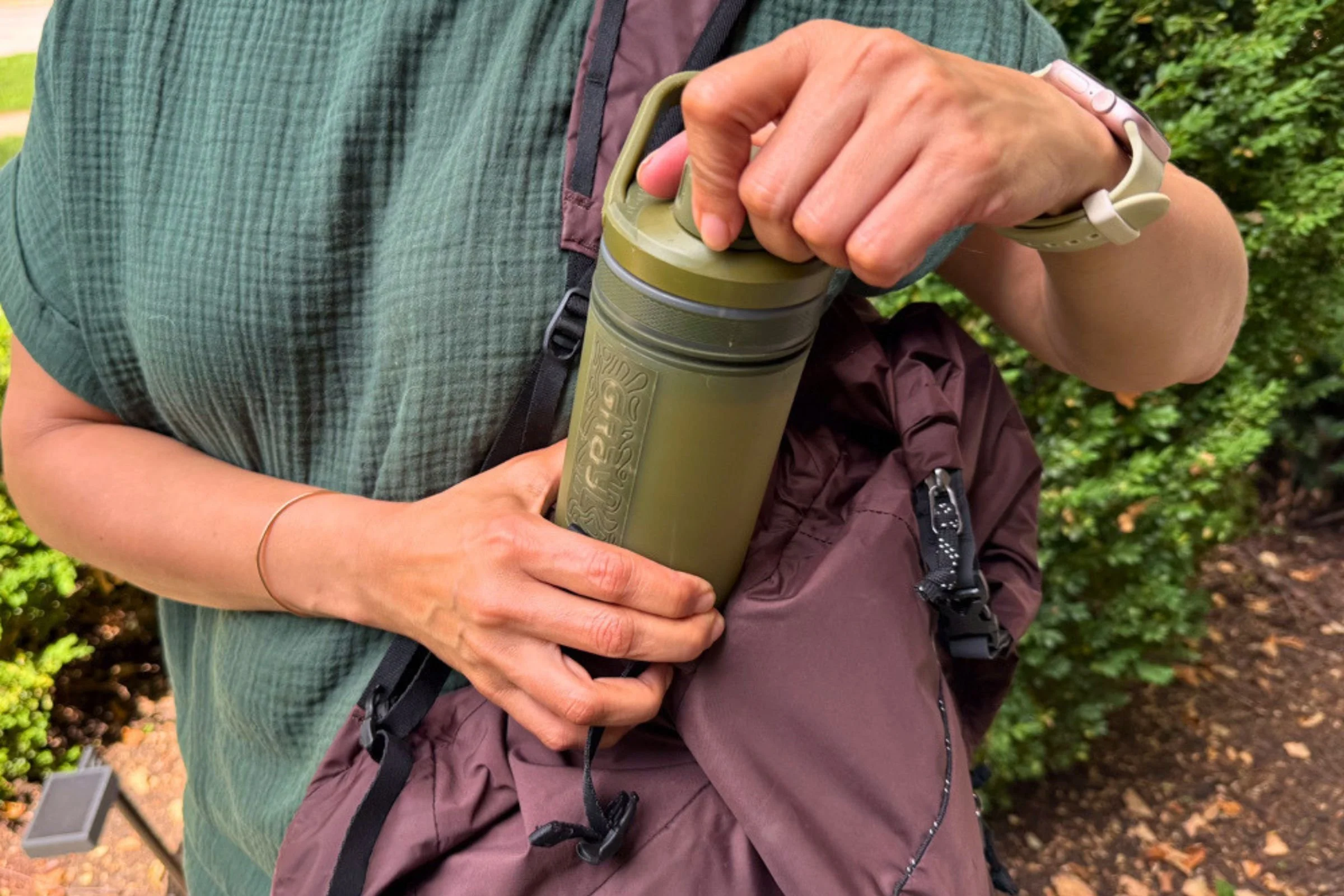 A person in a green shirt is placing Grayl UltraPress Filter and Purifier Bottle into a brown backpack.