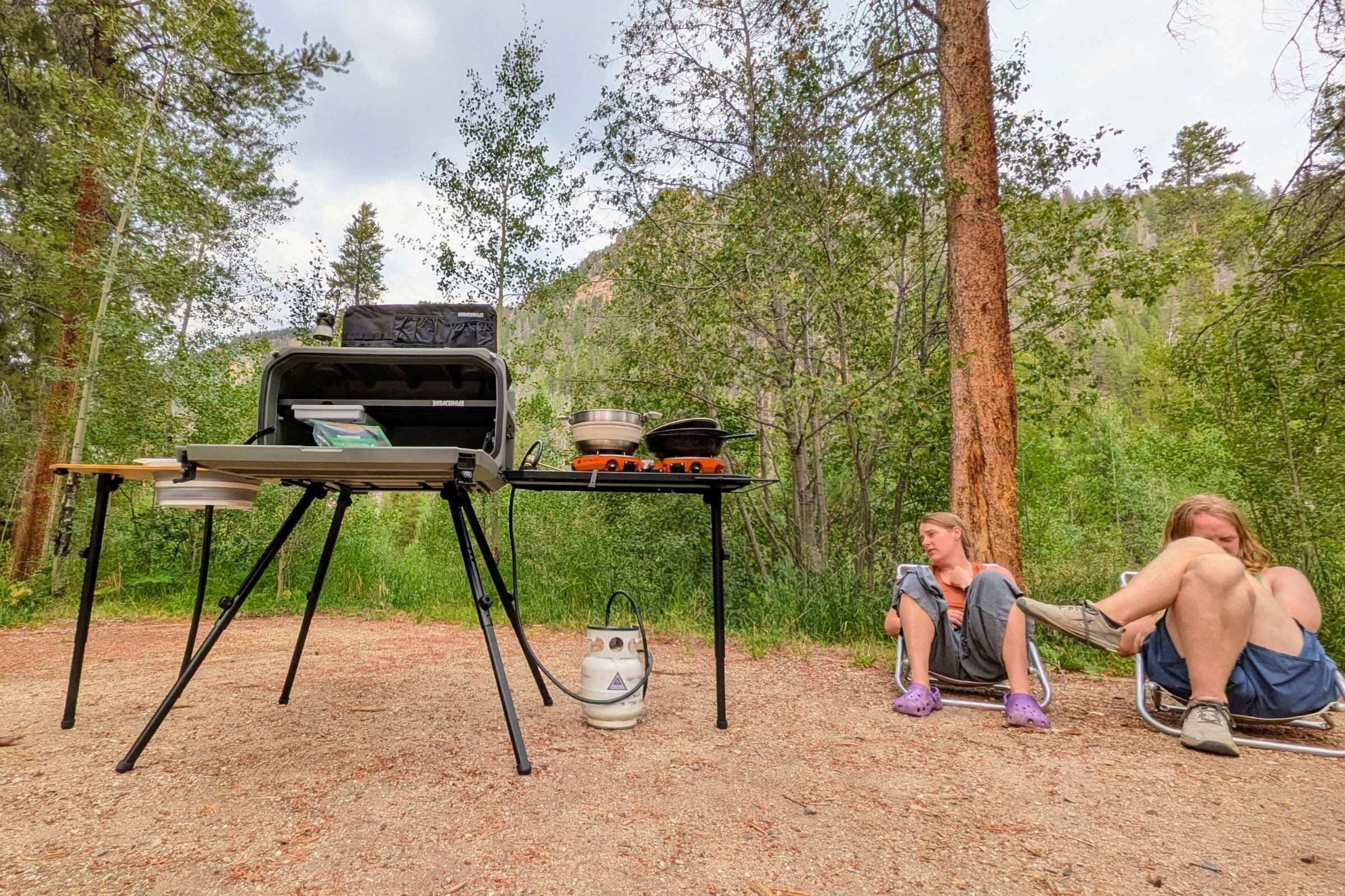 Two people seated in camp chairs with the Yakima EXO OpenRange freestanding leg kit nearby.