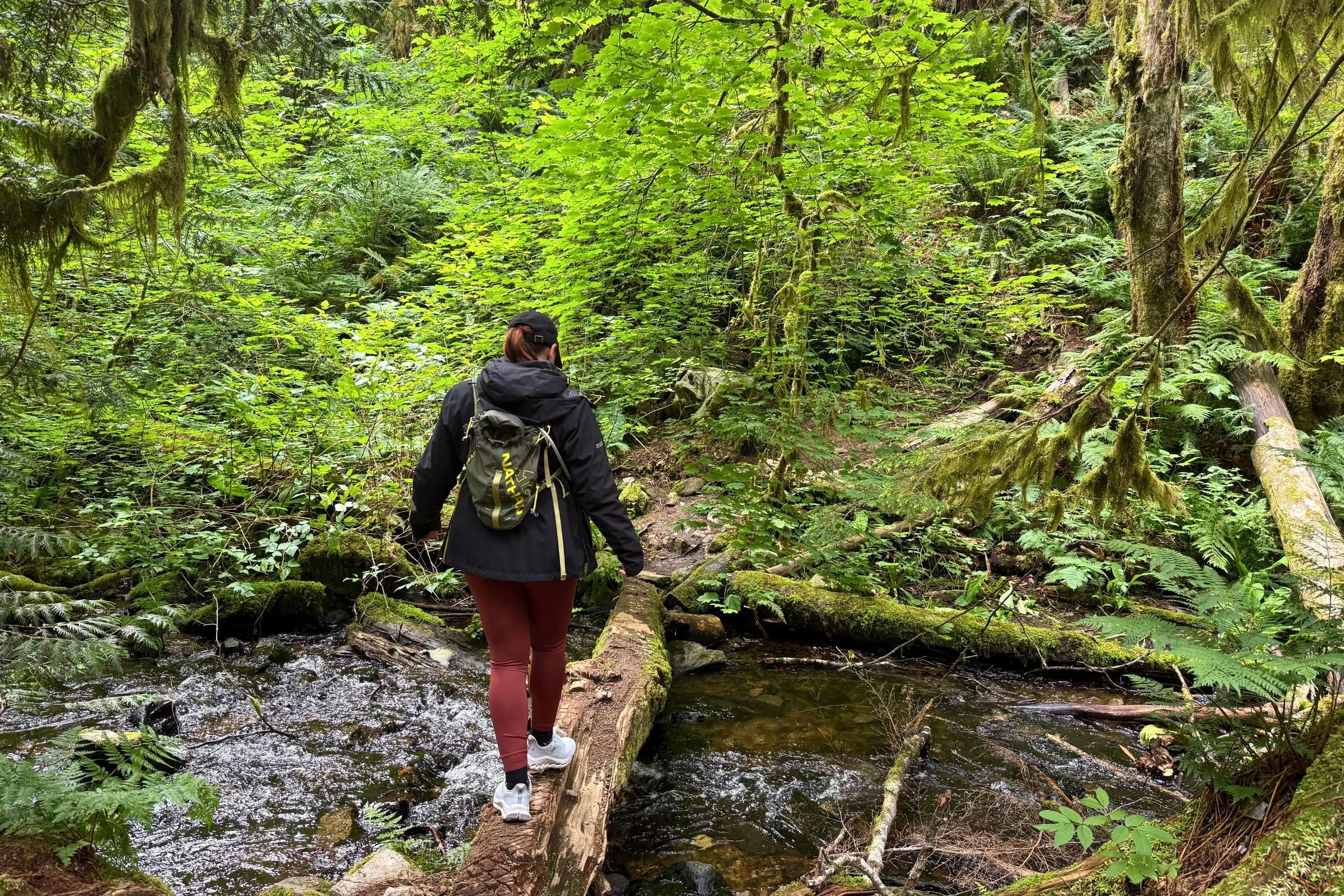 A woman wearing a backpacking while testing the grip of the On Cloudhorizon 2 hiking shoes on a wet log across a creek.