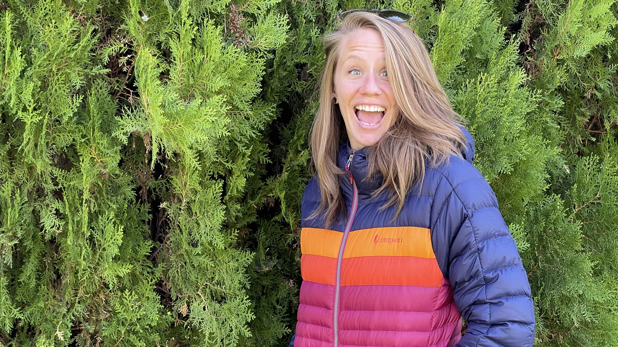the author wearing a colorful cotopaxi puffy jacket with green foliage in the background