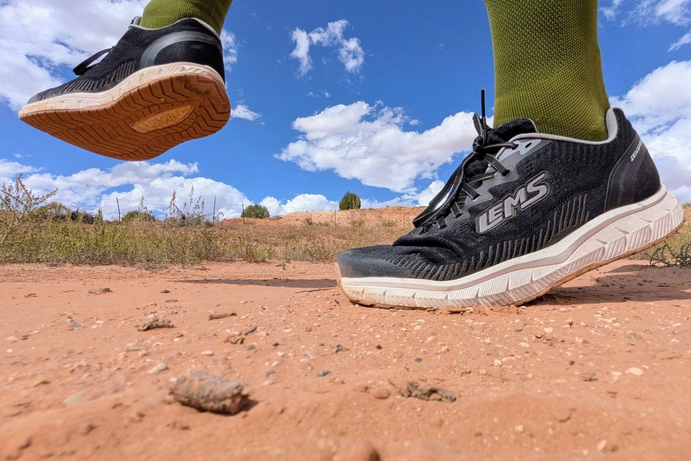 Person wearing black Lems ZENSPEED+ shoes while walking on a hard-packed dirt road.