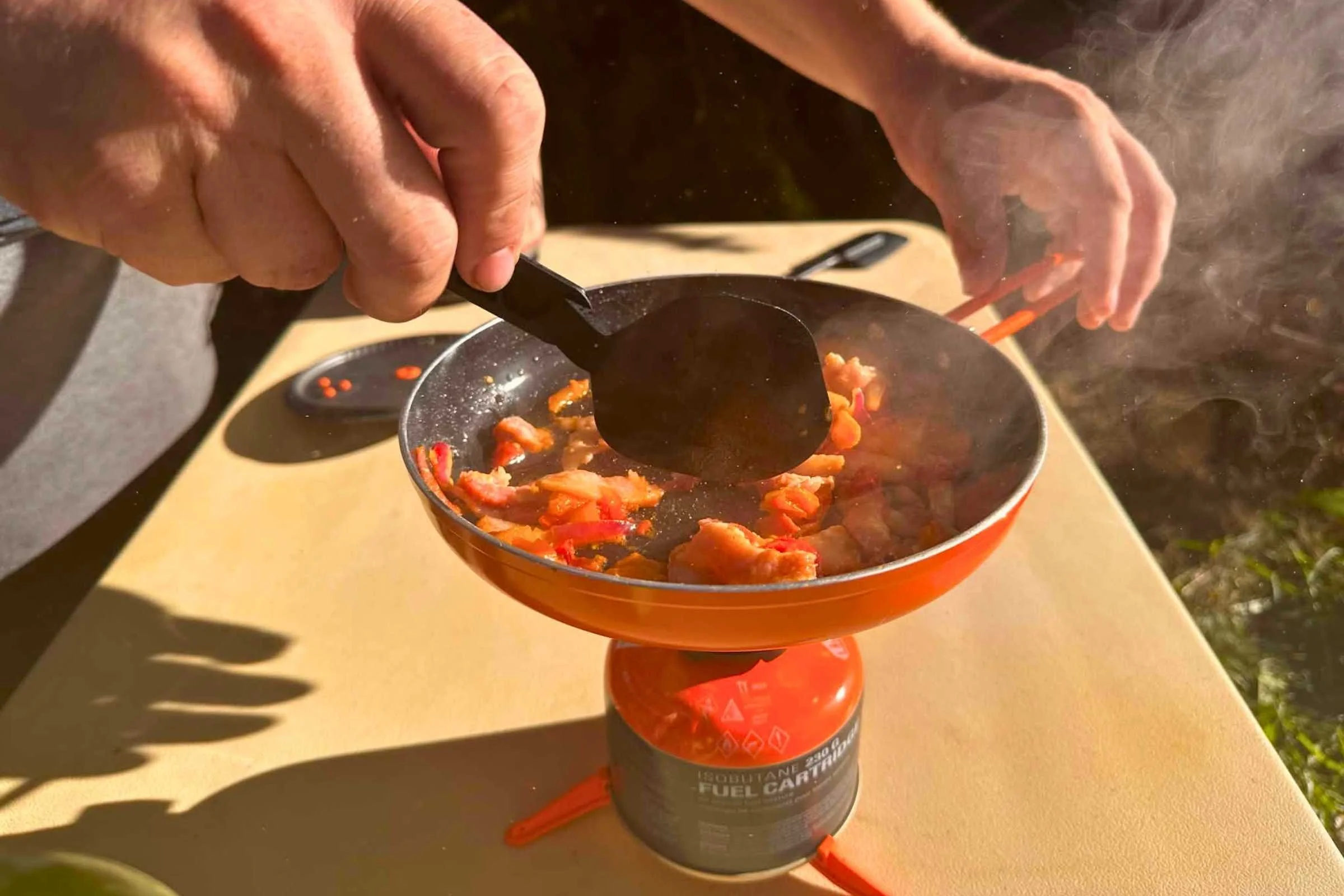 One hand holds the handle of the Jetboil Summit Skillet while another uses the included spatula to stir meat and vegetables as they cook on the TrailCook stove.