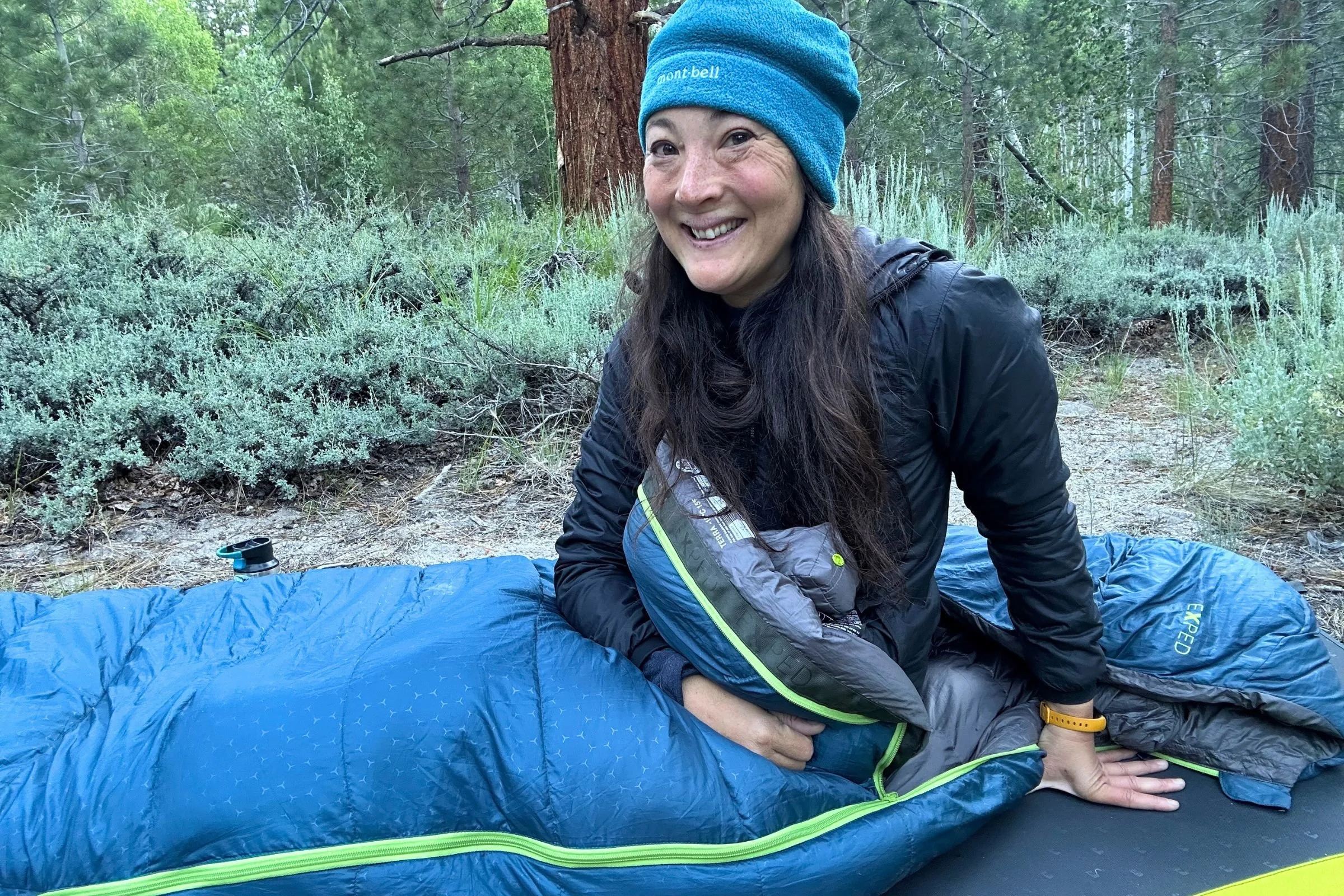A woman wearing a blue hat and blue jacket sits outdoors inside an Exped Terra Sleeping Bag.