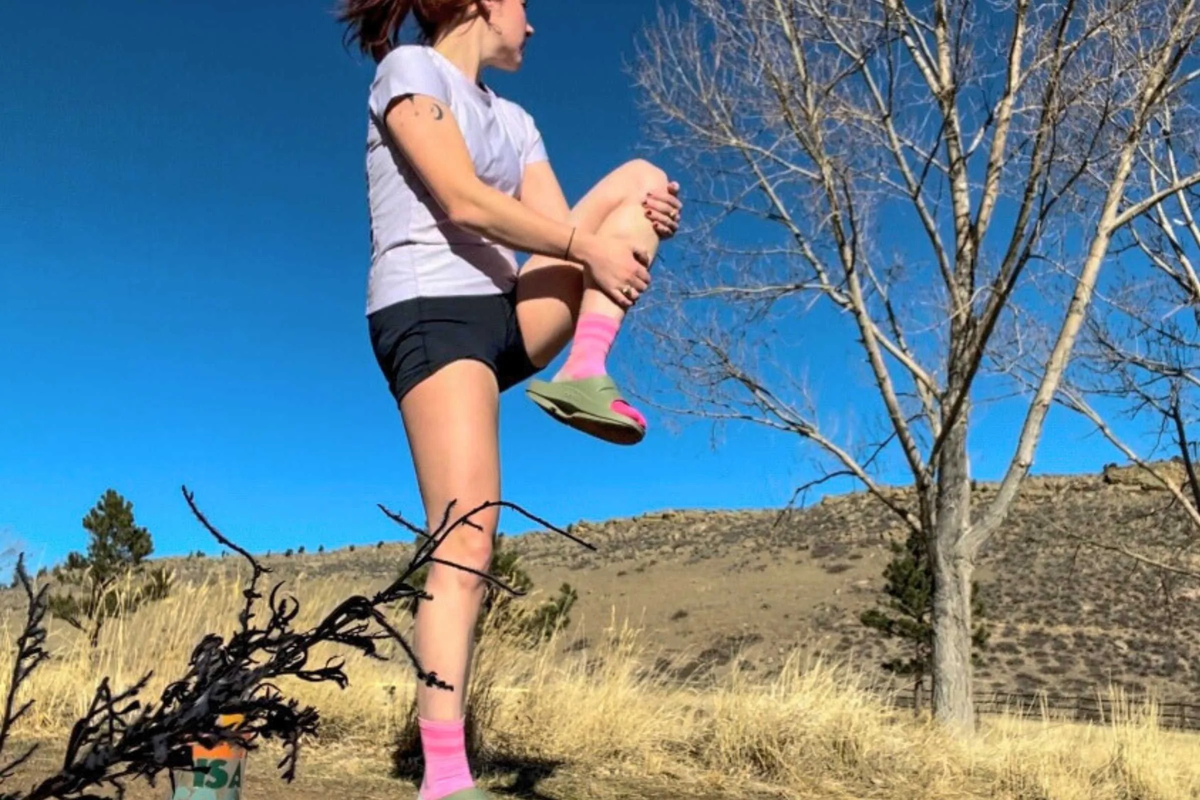 Person outdoors wearing OOFOS Ooahh recovery slides performs a standing knee stretch on grassy terrain