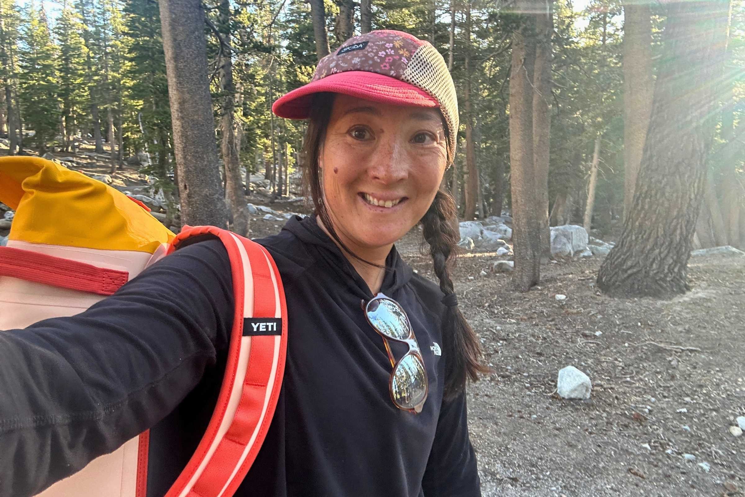 Woman wearing The North Face Sunriser Sun Hoodie while carrying a backpack in a forest campsite