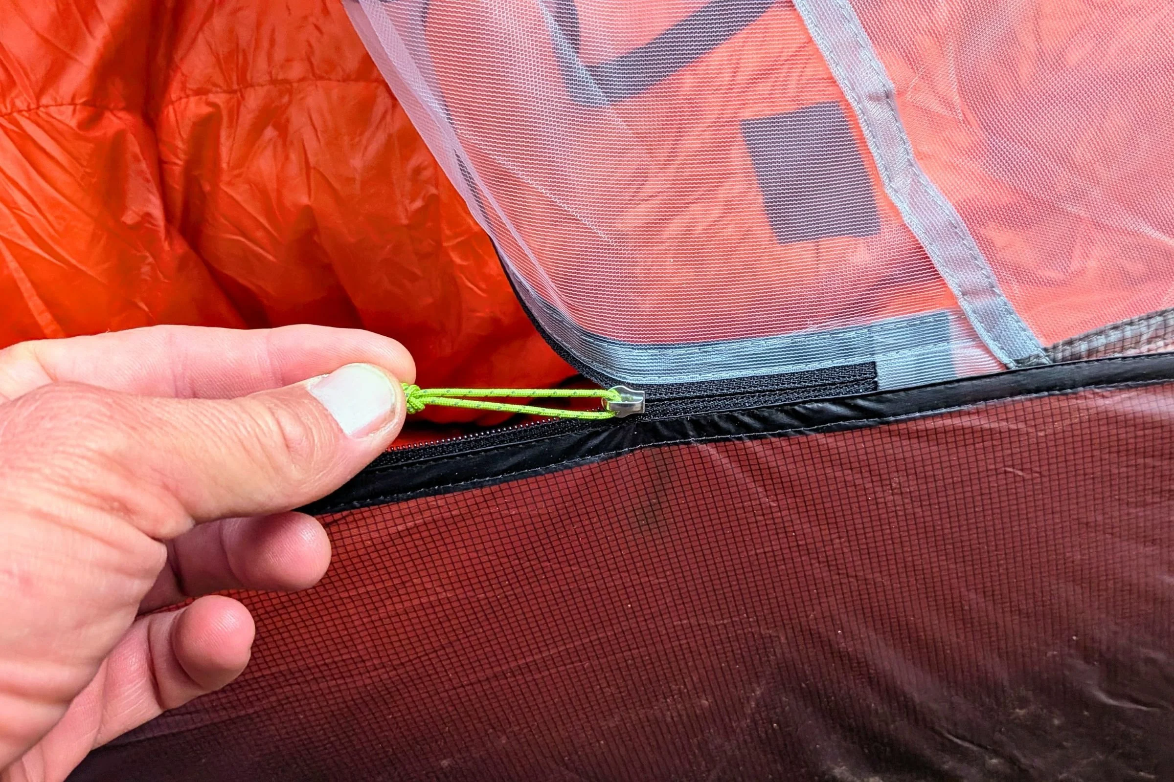 A hand holds the inner-door zipper pull of Gossamer Gear The Free beside a sleeping bag.