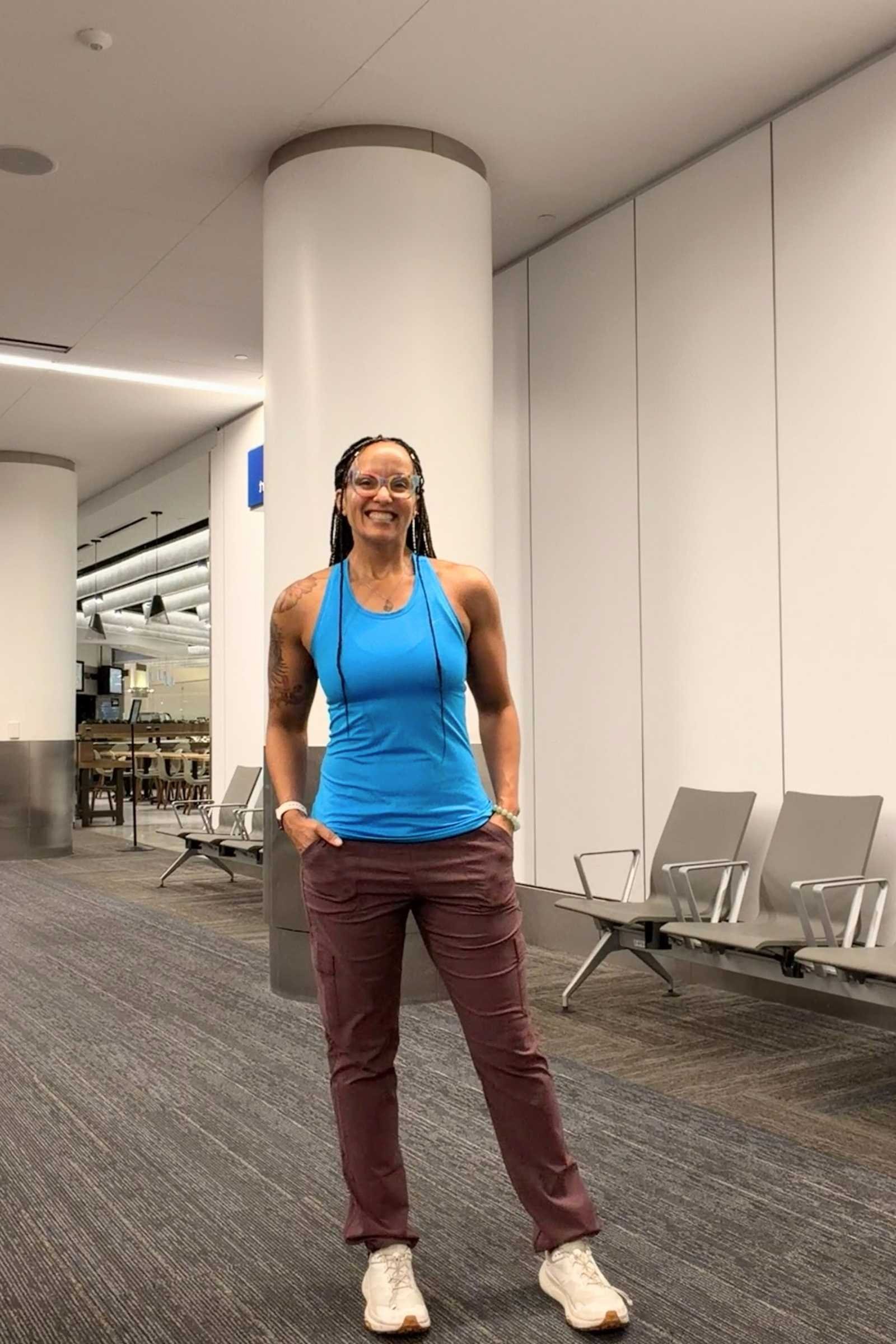 Woman wearing Kuhl Trekr Straight travel pants in raisin color and blue tanktop in an airport (a city environment)
