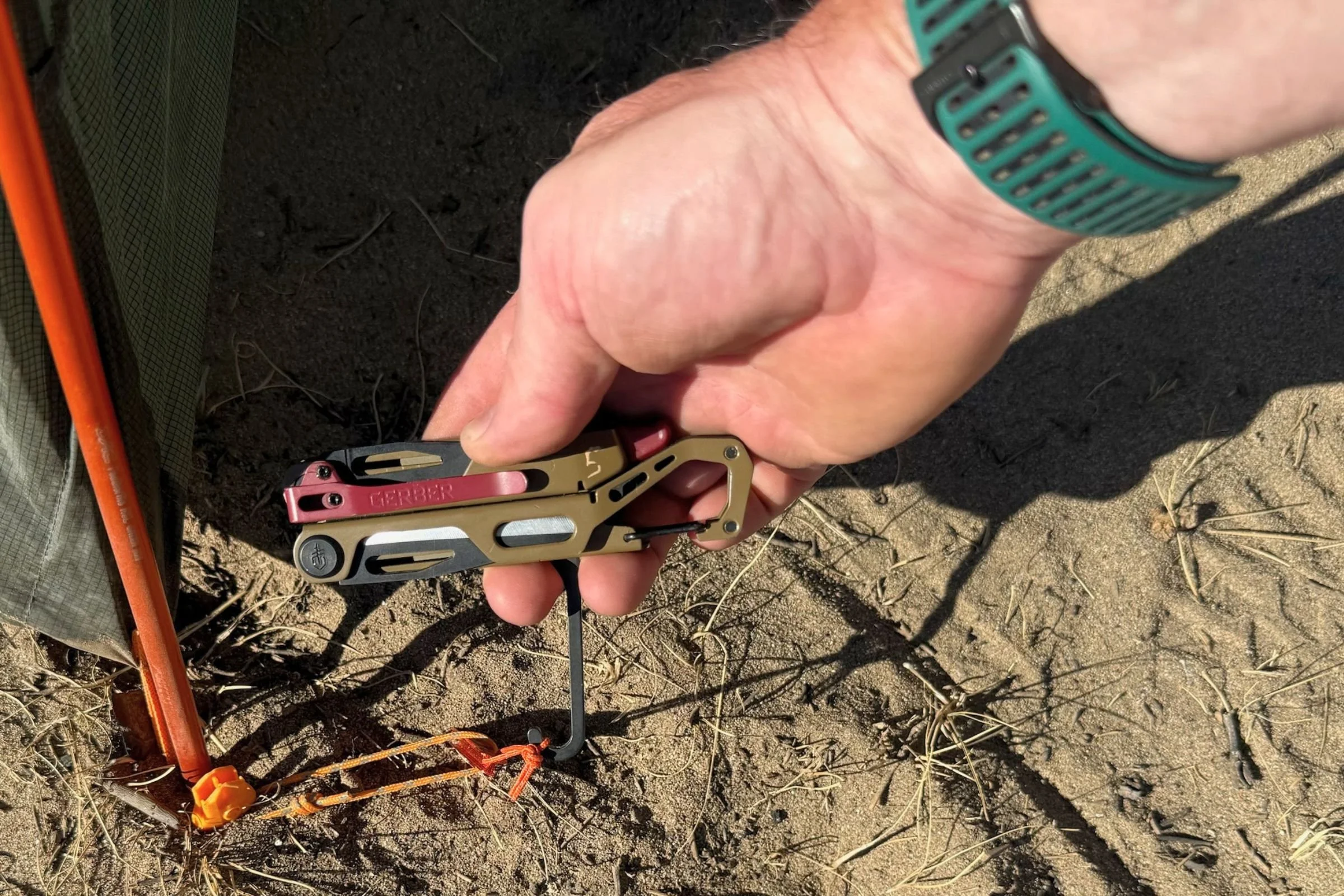 Using the Gerber Stakeout Spark’s hook to pull a tent stake from sandy ground beside a tent.