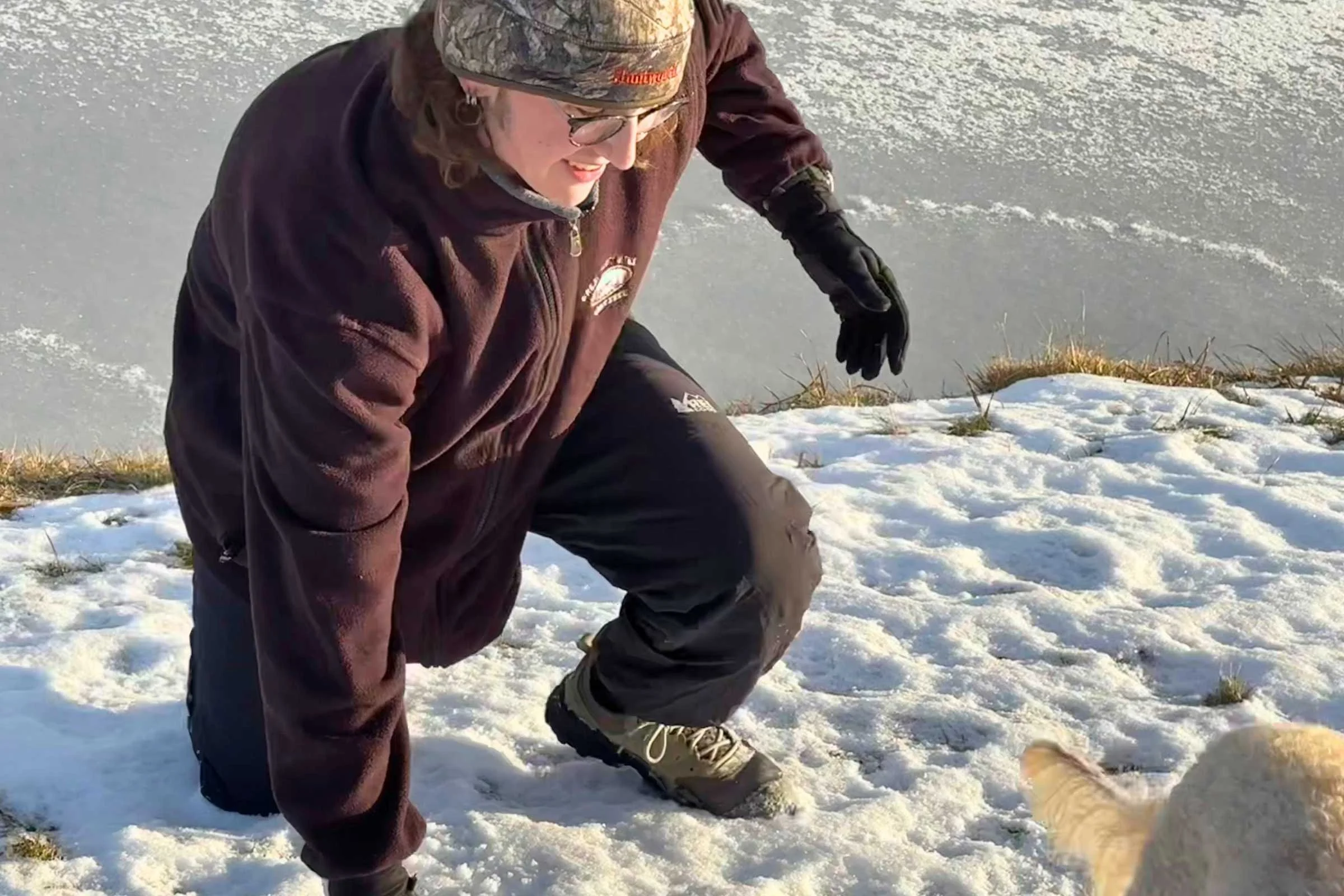 Elliot is kneeling in the snow in the REI Co-op XeroCloud rain pants with a dog in the corner of the frame.