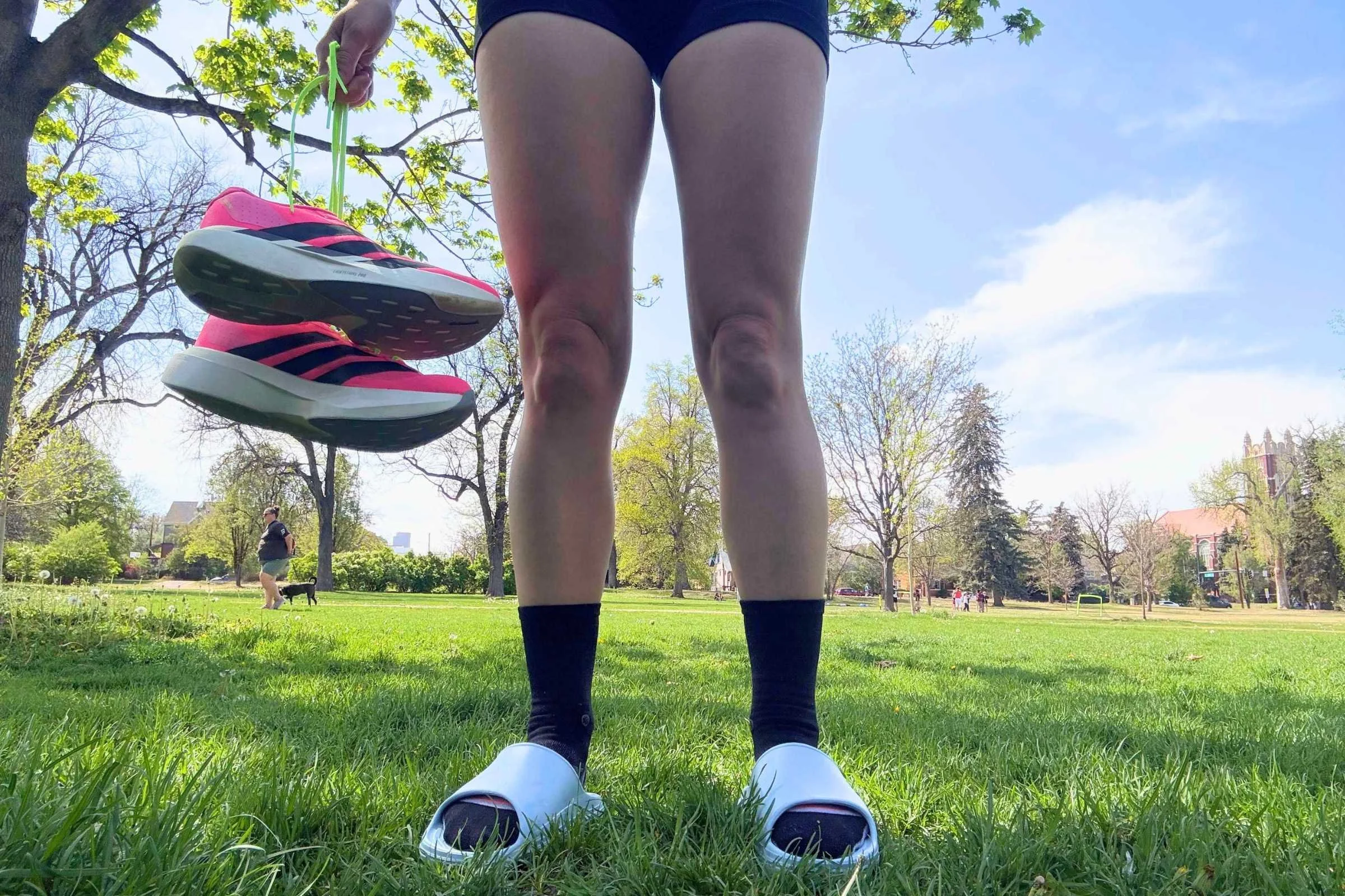 Norda 008 Recovery Slides: A woman in shorts standing in the grass holding a pink running shoe and wearing recovery slides.