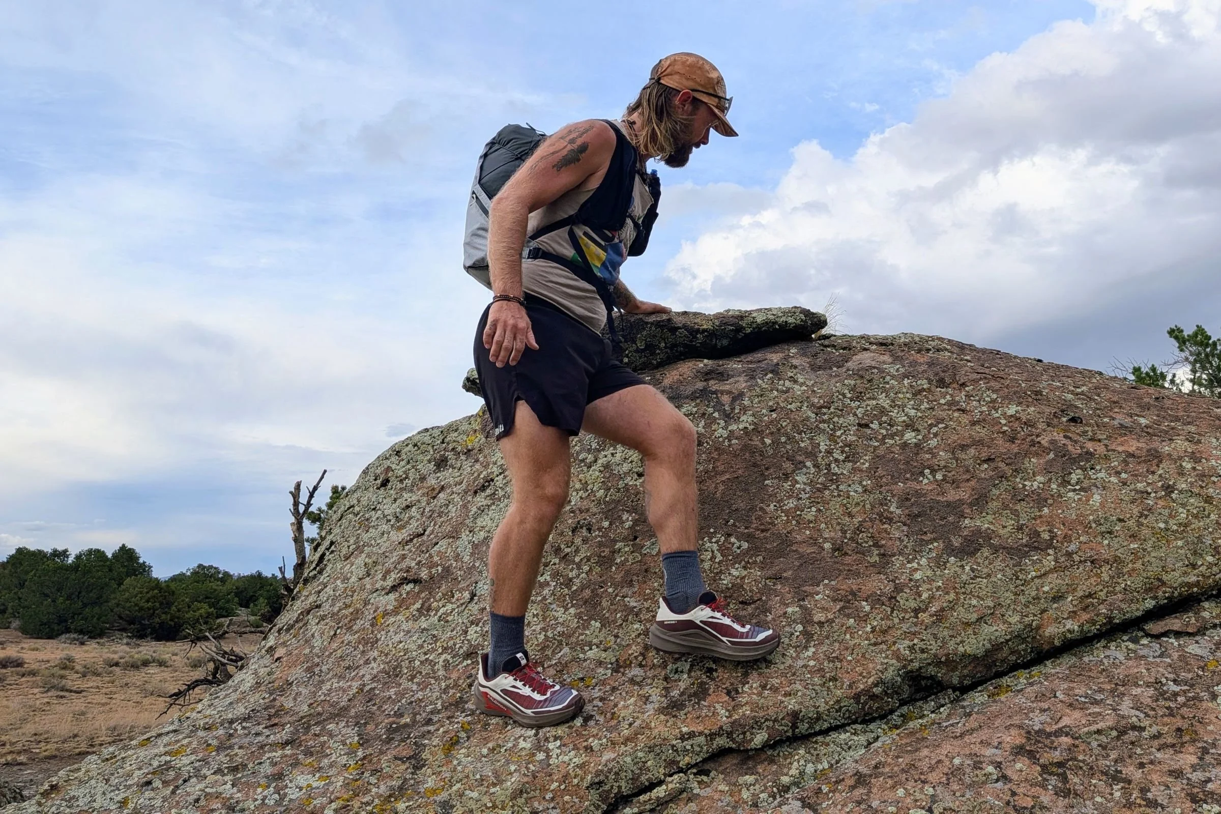 A man in a hat and shorts wearing Salomon Genesis 2 Trail Running Shoes climbs a rock.