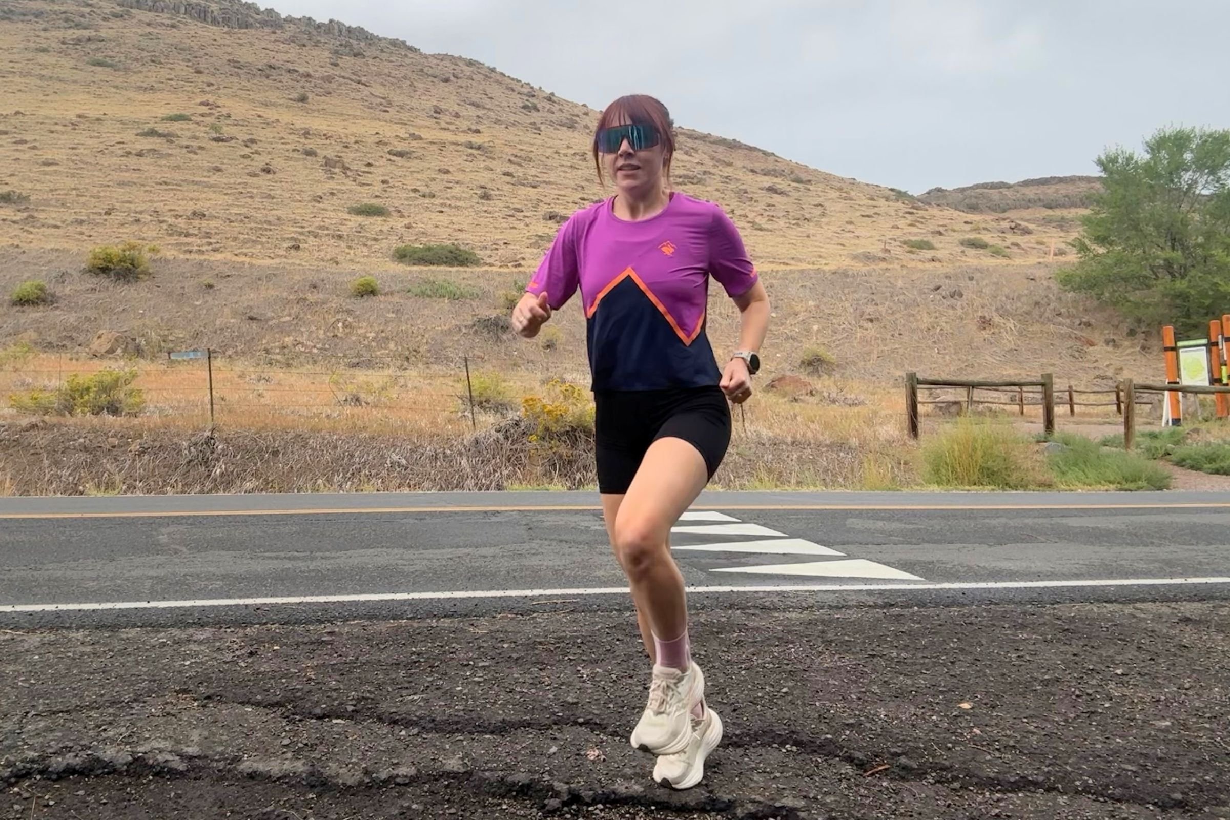 A woman in a purple shirt and shorts running on a road in the Saucony Triumph 24 Road Running Shoe.