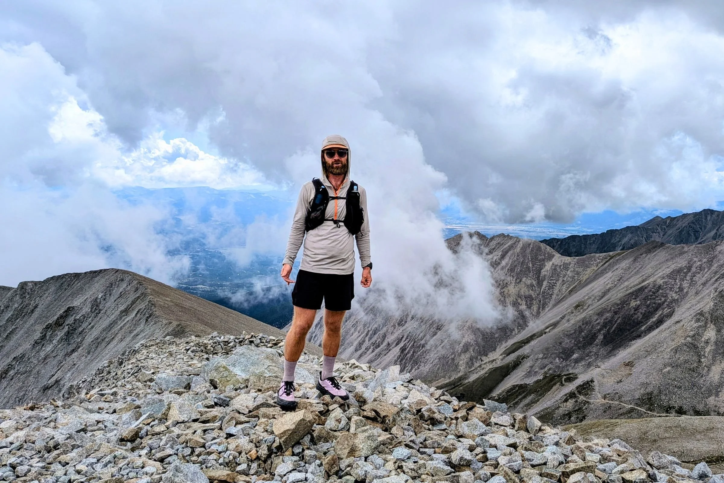 Man standing on a mountain summit with clouds in the background, wearing Norda 000 Trail Running Shoes.