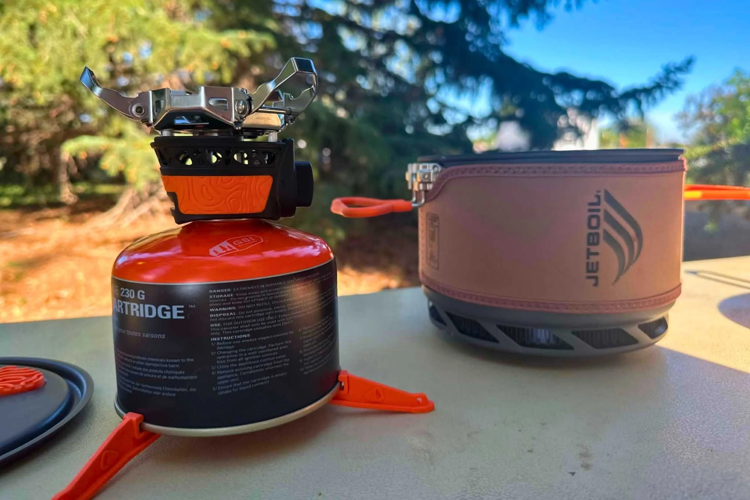 The Jetboil TrailCook burner attached to an isobutane/propane canister and plastic base with the pot and pot lid on either side.