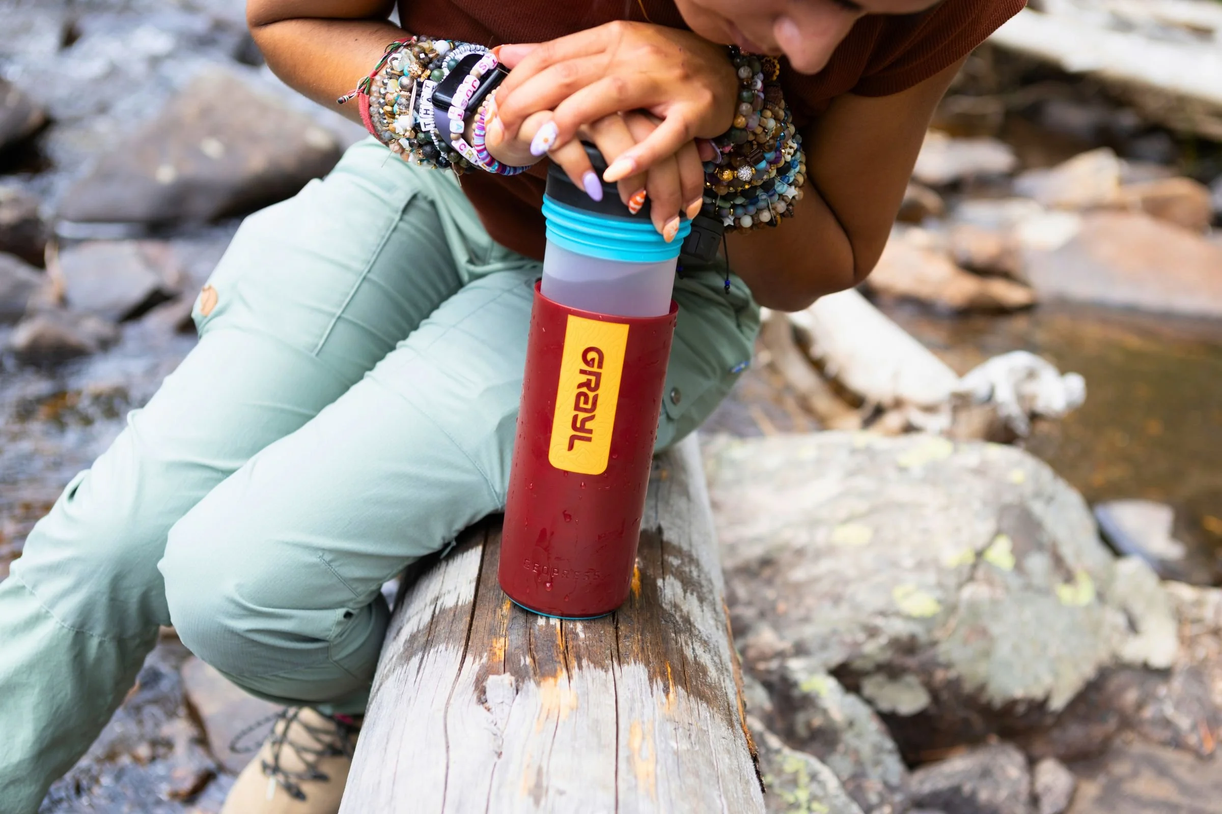 A woman sitting on a log while holding a Grayl GeoPress Purifier.