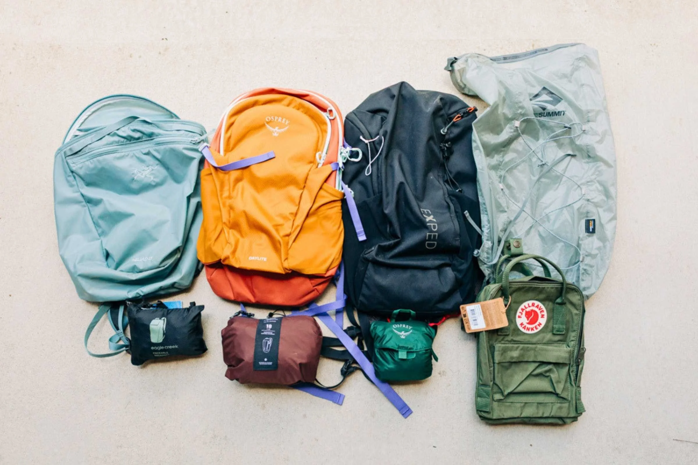 Eight packable backpacks laid flat on a neutral surface