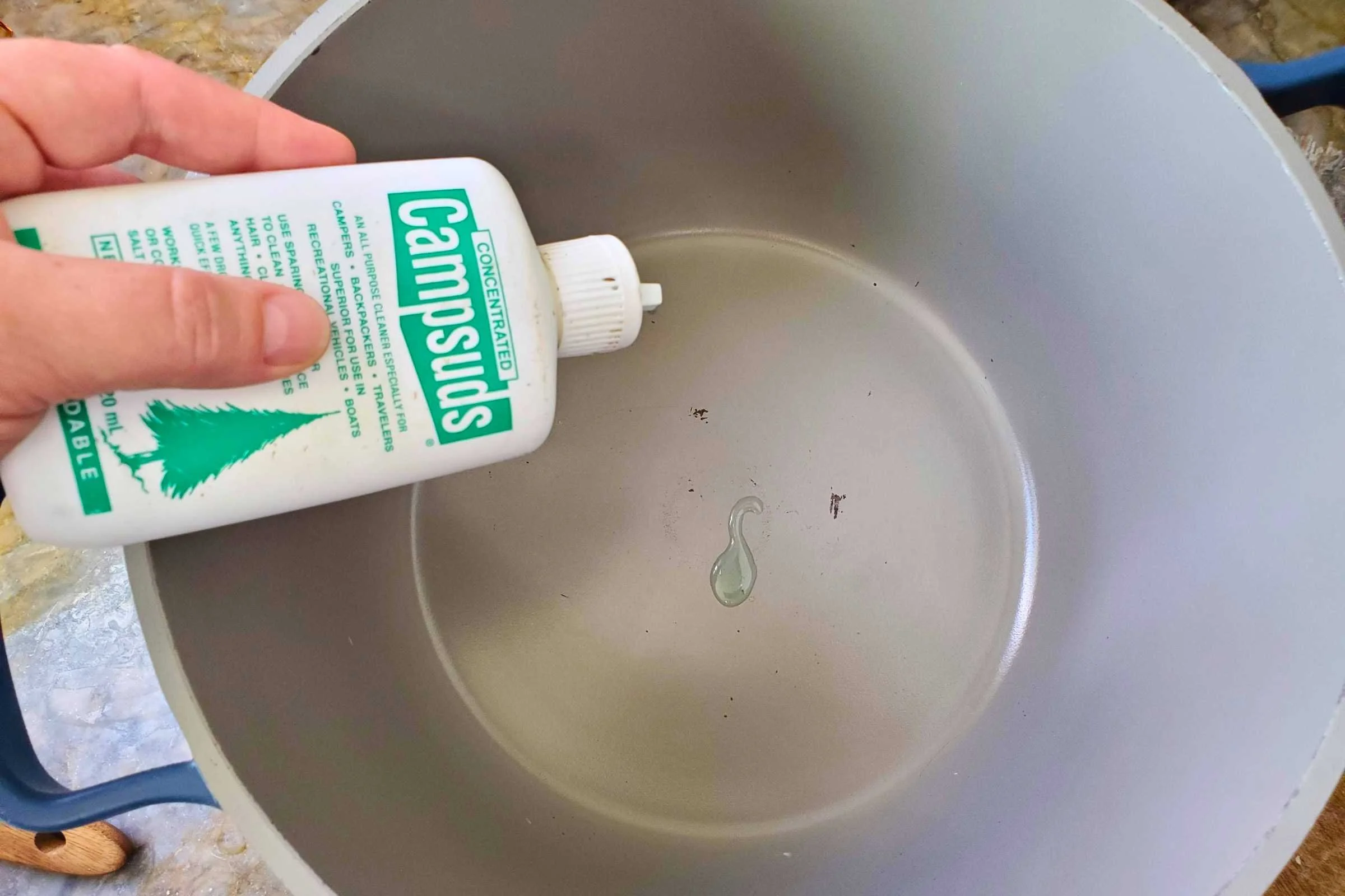 A hand holds Campsuds Biodegradable Soap over a large pot, where a few drops of soap are visible in the bottom