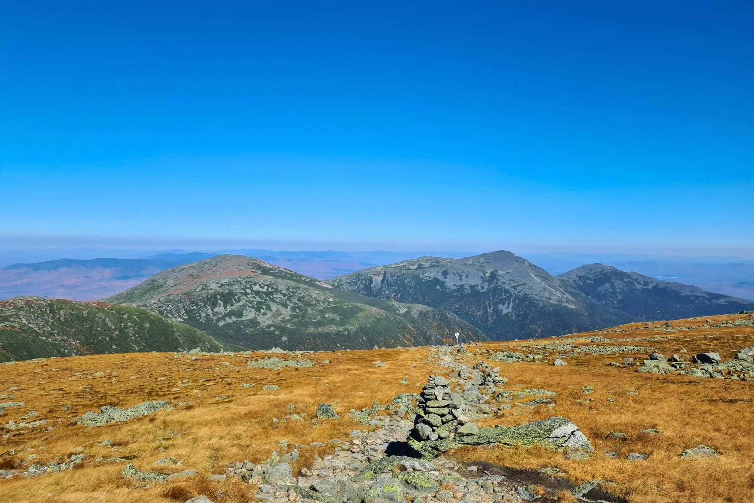 Cairns on the Appalachian Trail with a trail sign in the distance, and Mt. Clay, Jefferson, Adams, and Madison in the background. Photo by Aubri Drake.