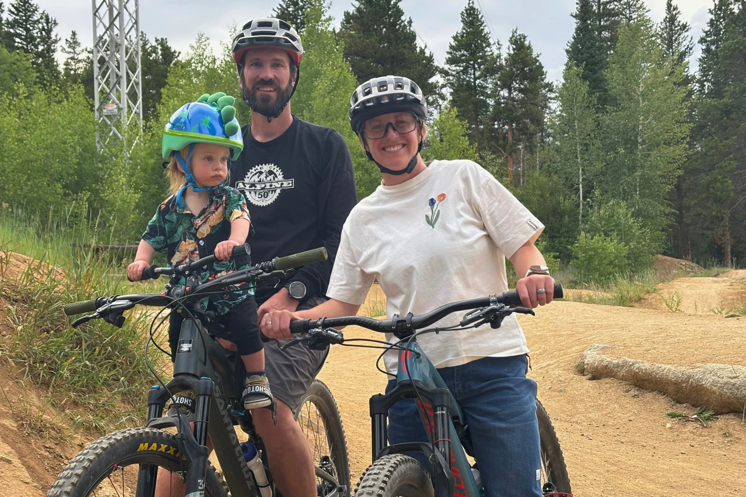 Father and son on a Santa Cruz mountain bike, with the child in the Kids Ride Shotgun Shotgun Pro EVO 2-in-1 bike seat, mother on her own bike, with trees and MTB trails in the background.