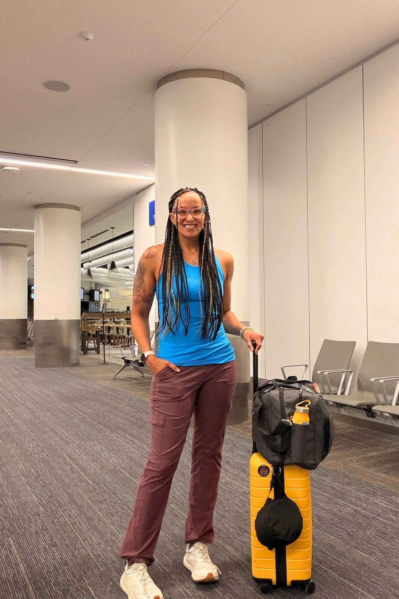 A person walking through an airport terminal wearing Kuhl Trekr Straigh travel pants in raisin color with a carry-on bag and personal item