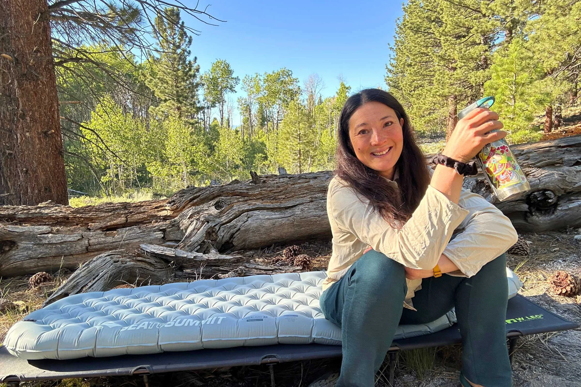 Alpenblow Lux camping mat scene with a woman sitting beside a water bottle outdoors.