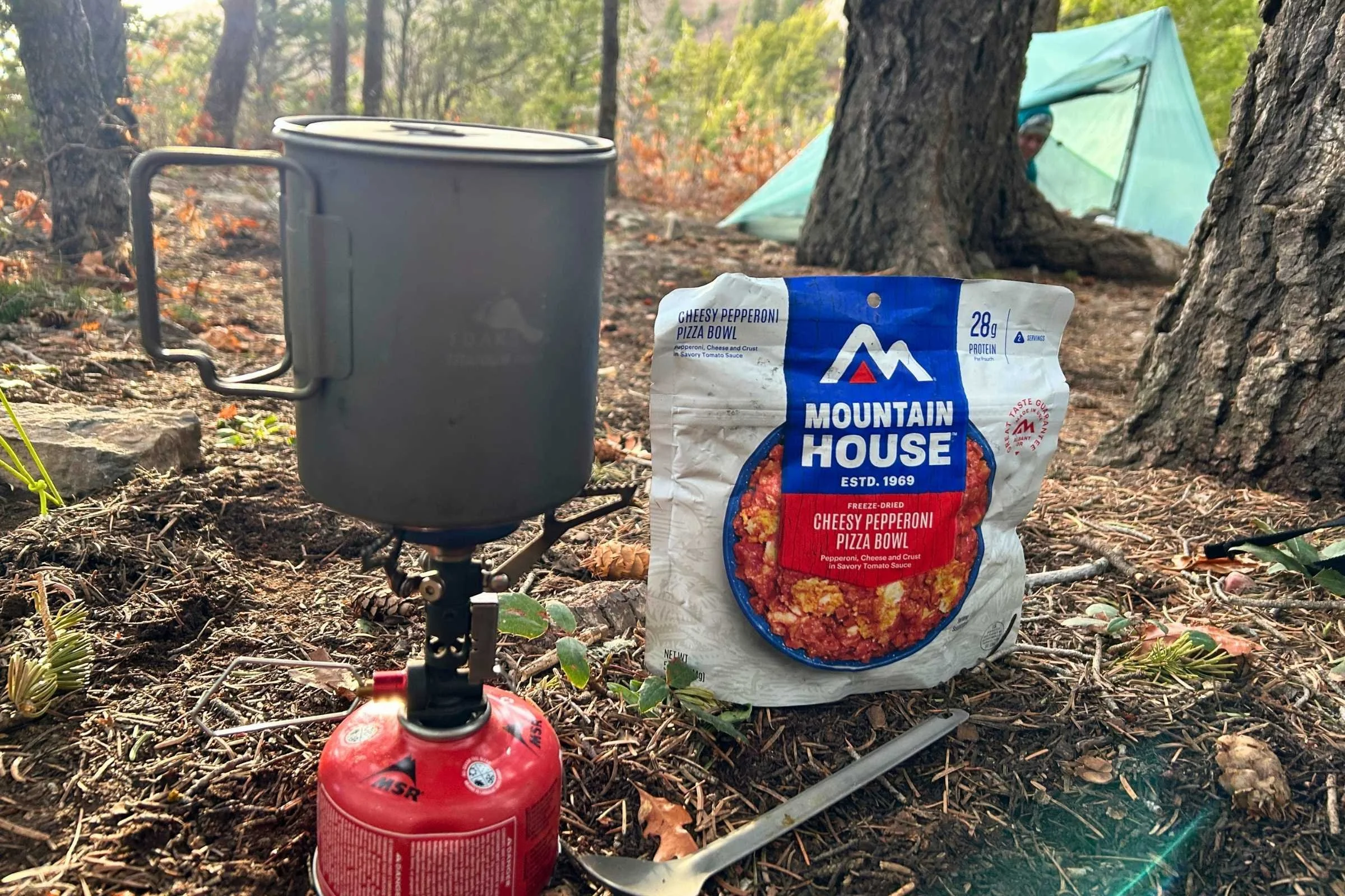 Mountain House Cheesy Pepperoni Pizza Bowl freeze-dried meal next to a backpacking stove and pot at a forest campsite.