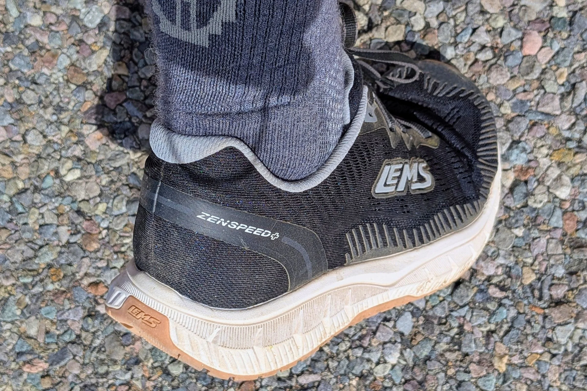 Person wearing black Lems ZENSPEED+ shoes while standing on a gravel road.