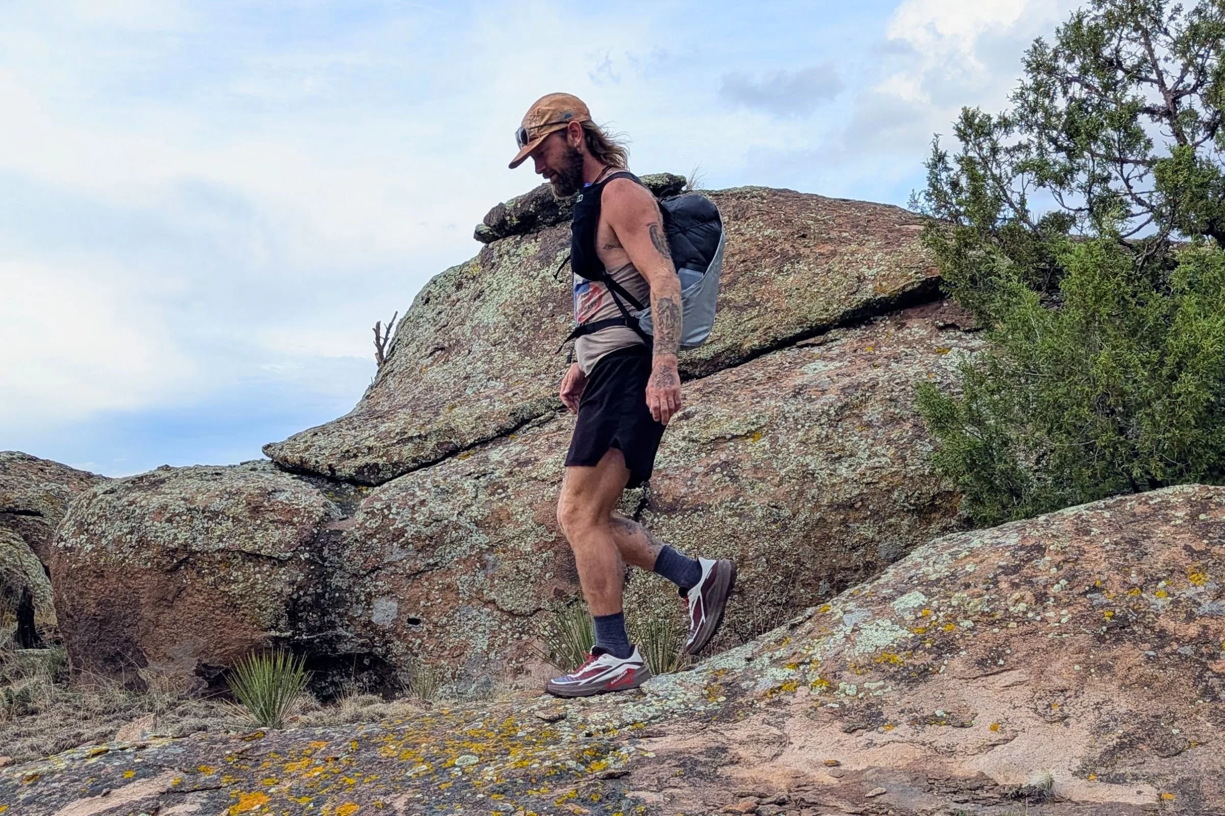 A man with a backpack and hat wearing Salomon Genesis 2 Trail Running Shoes walks across rocky terrain.